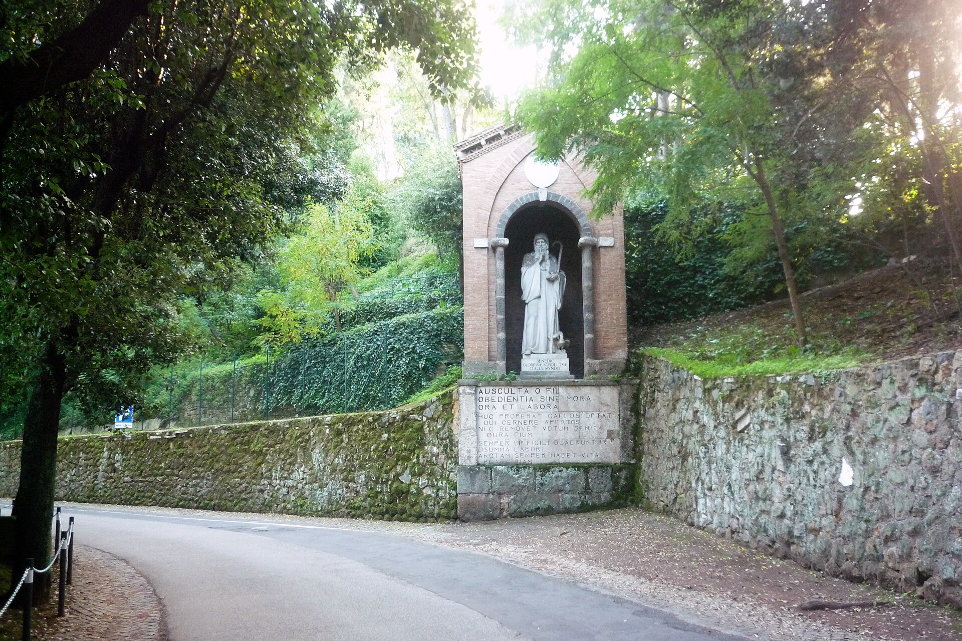 Photograph of a group of old brick and stone church buildings among trees, with a gateway in the foreground