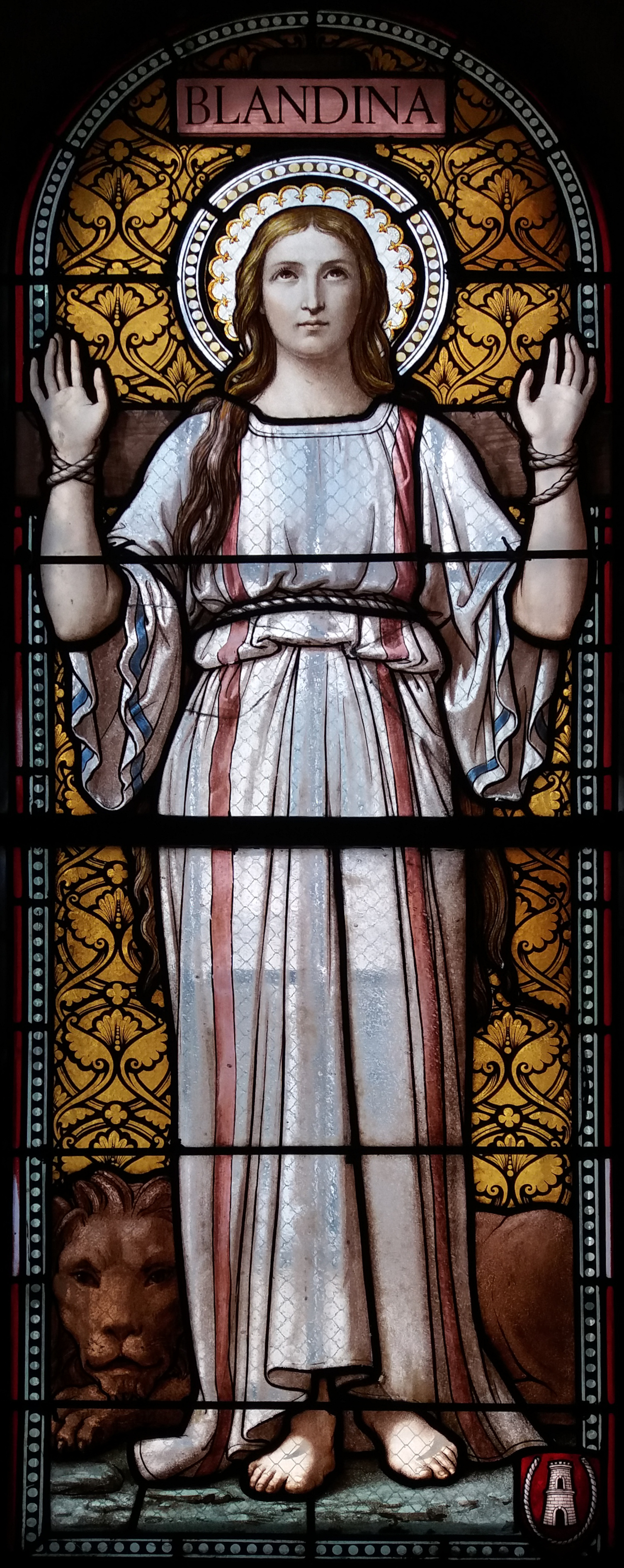 Tall stained glass window showing a young woman in a long robe with a halo, standing with her hands joined, a lion at her feet