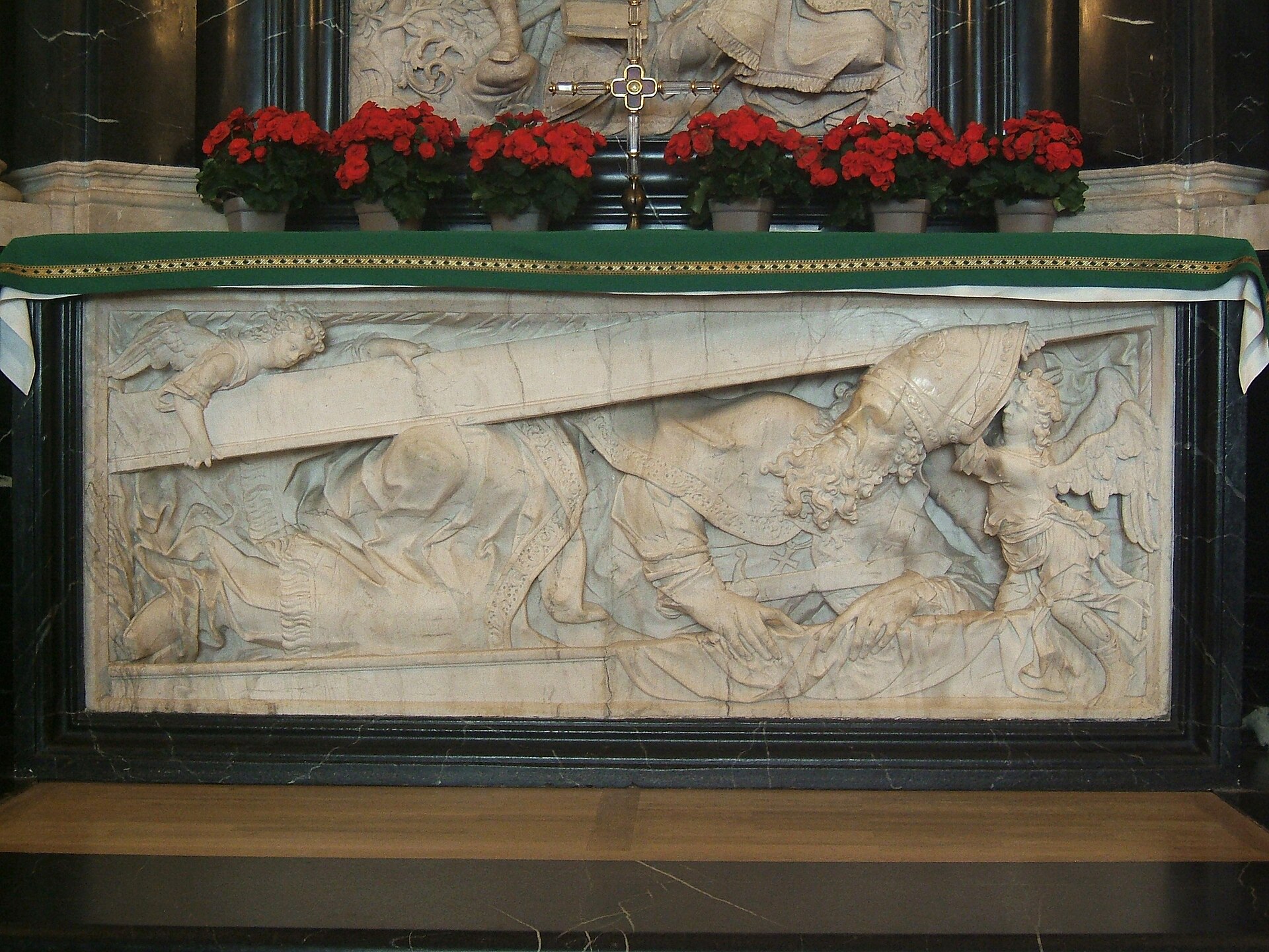 A white carved relief panel on the front of an altar in the crypt of Fulda Cathedral, with flowers set above it