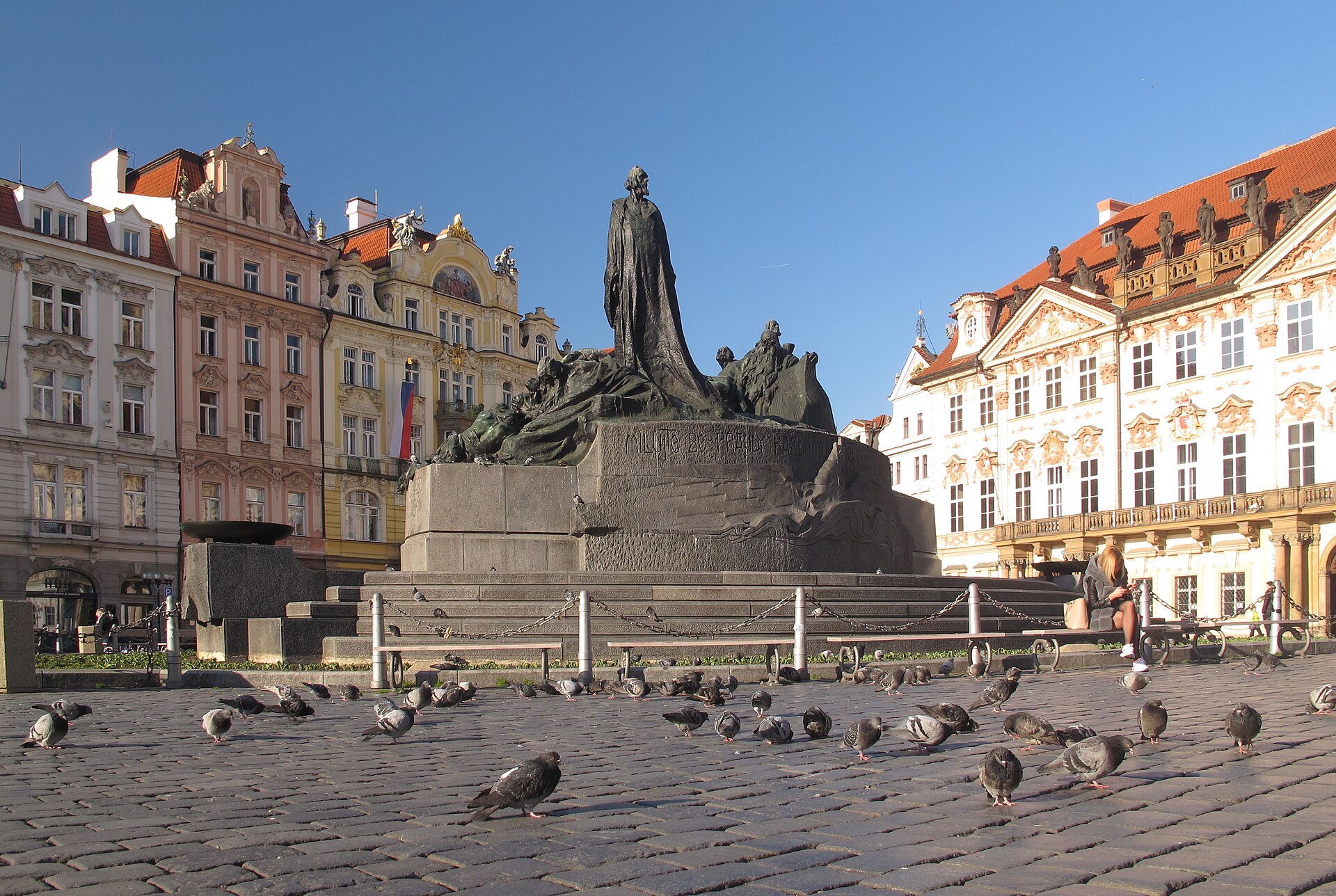 Large bronze monument of a tall robed figure standing above groups of smaller figures on a stone base in an almost empty city square
