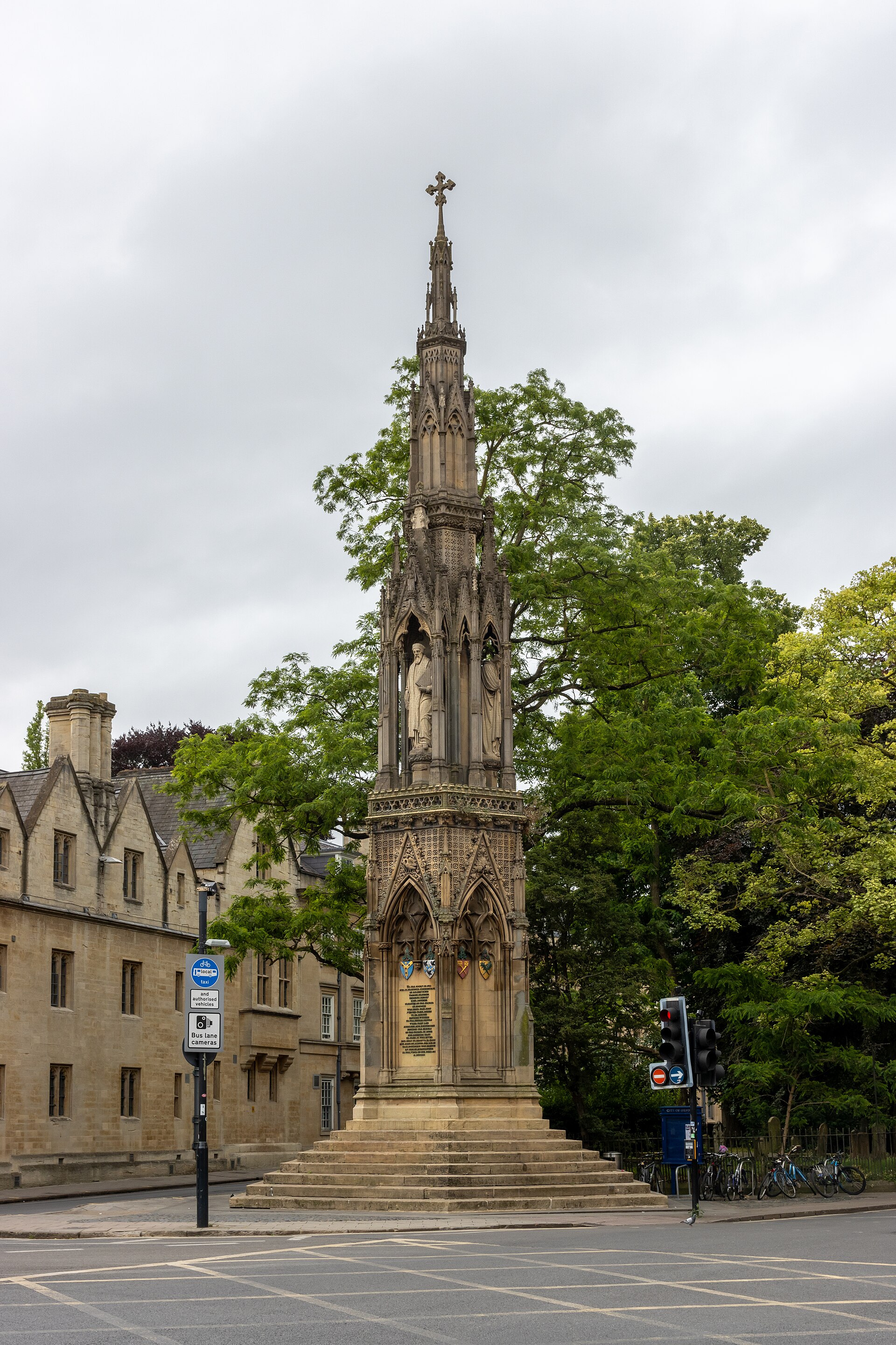 A tall carved stone Gothic monument with statues in niches, standing at a road junction in Oxford under a clear sky