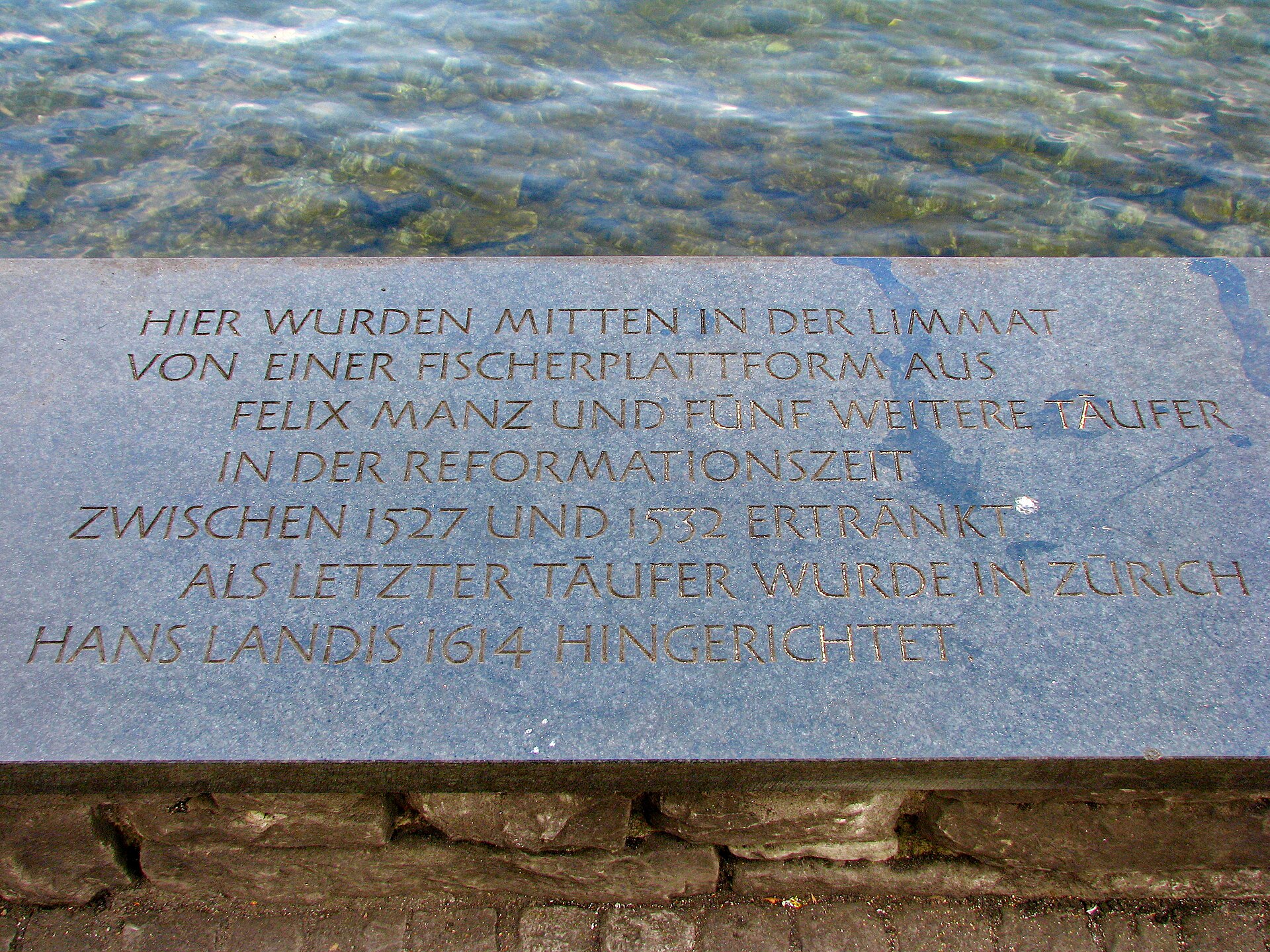 A dark stone plaque with carved German lettering set into a river embankment wall in Zurich
