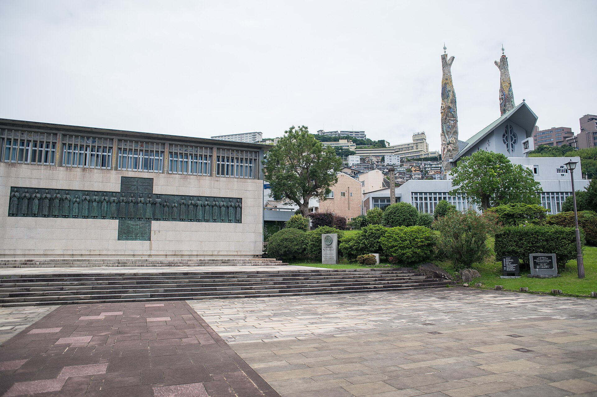 A low modern stone building with a long dark panel across its front, a paved plaza with steps and shrubs, and two tall slender church spires rising behind trees