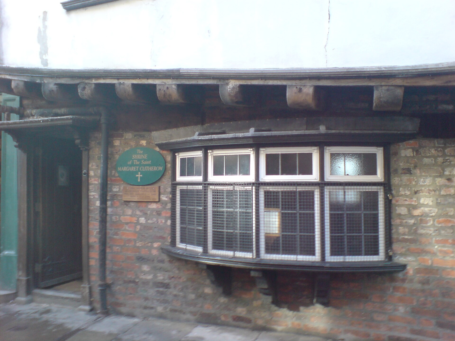 A narrow brick shopfront with a wide multi-paned bay window under a timber overhang and a round green plaque beside a dark doorway