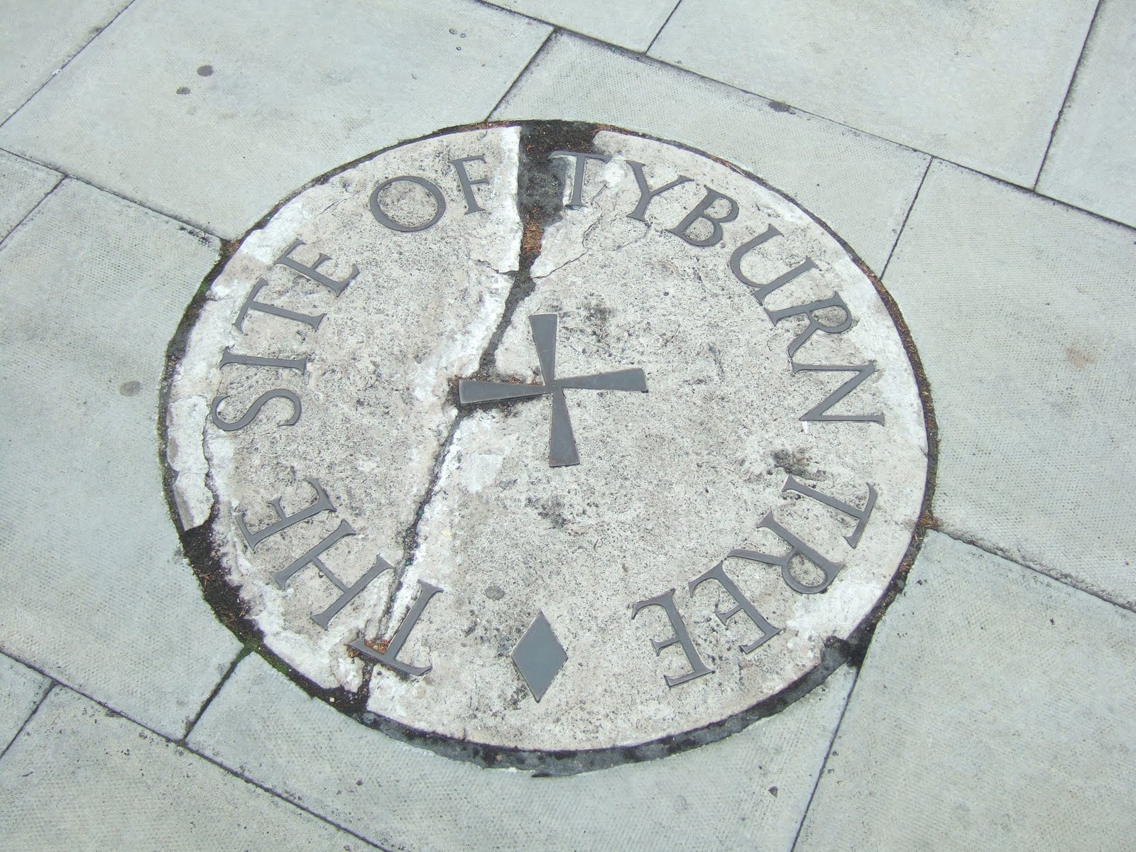 A round stone set into gray paving slabs, inscribed THE SITE OF TYBURN TREE around a small cross, with a crack running across it