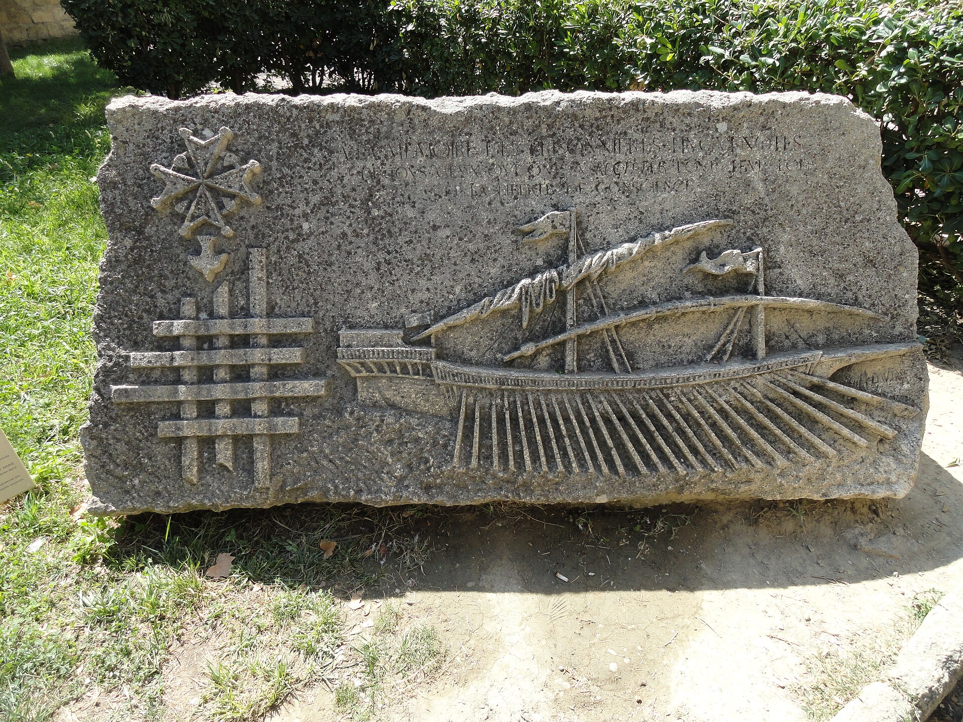 A carved stone memorial plaque on a courtyard wall, showing a Huguenot cross above a grille of bars.