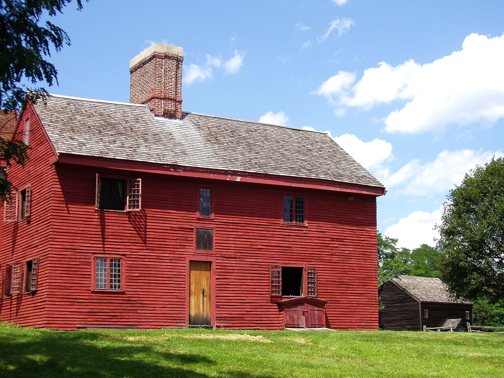 A dark brown seventeenth century timber farmhouse with a steep roof and a central chimney, standing in a grassy yard under trees.