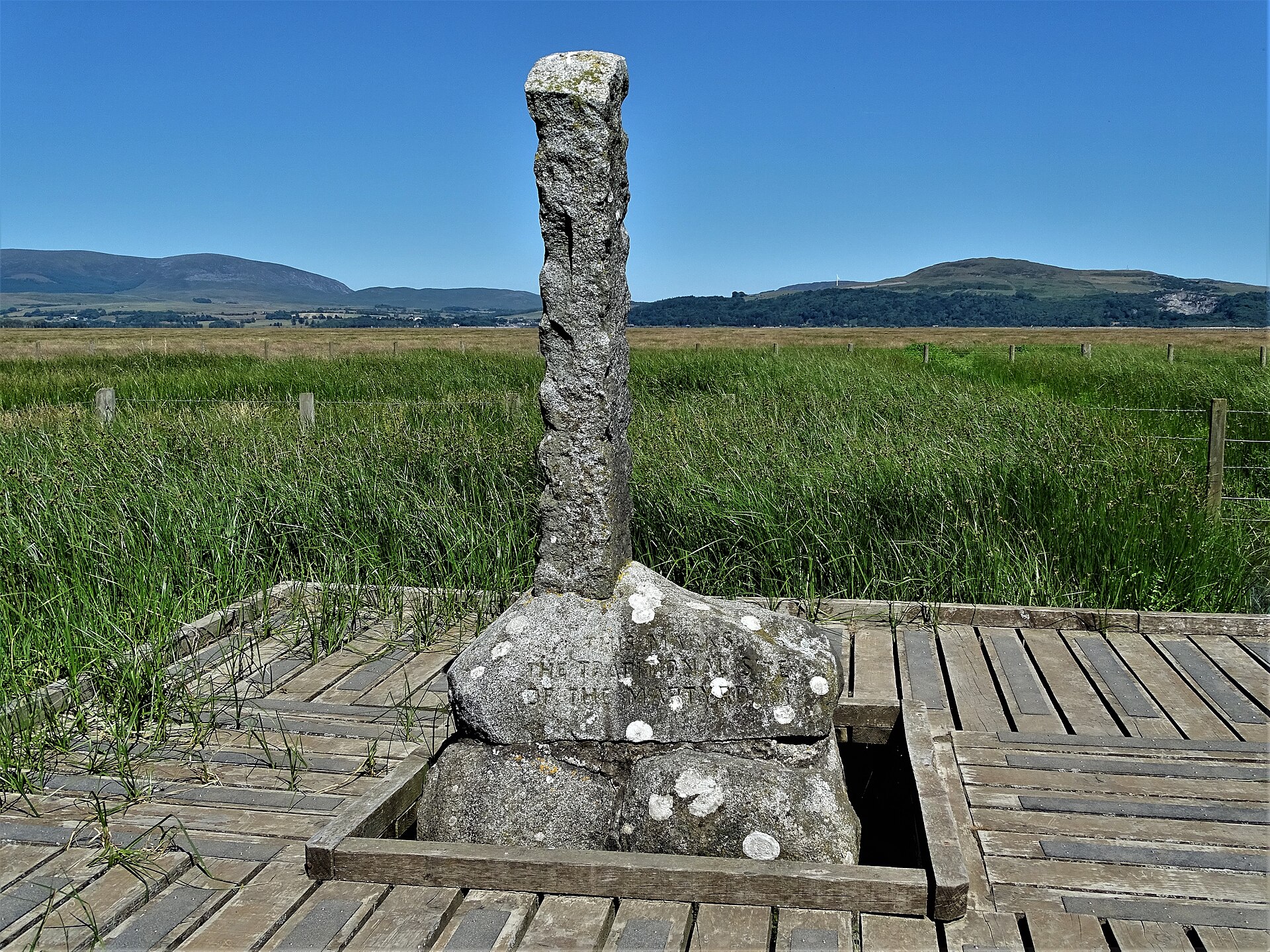 A wooden post standing in a salt marsh with a stone platform and railing in front of it, a modern marker at the place where the Wigtown martyrs are said to have died.
