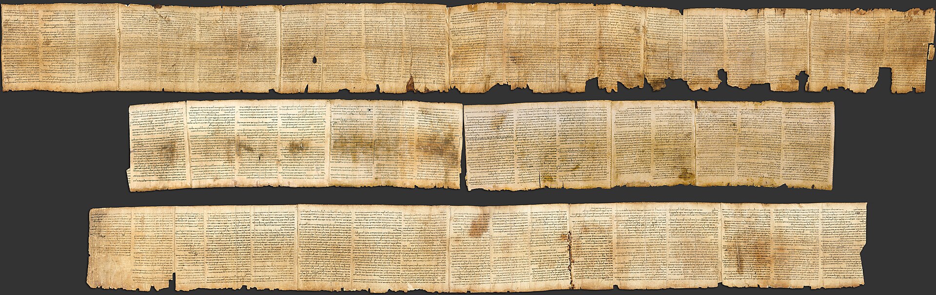 A long, narrow photograph of an unrolled, yellowed scroll showing many columns of dense Hebrew handwriting, with darker worn patches and small tears