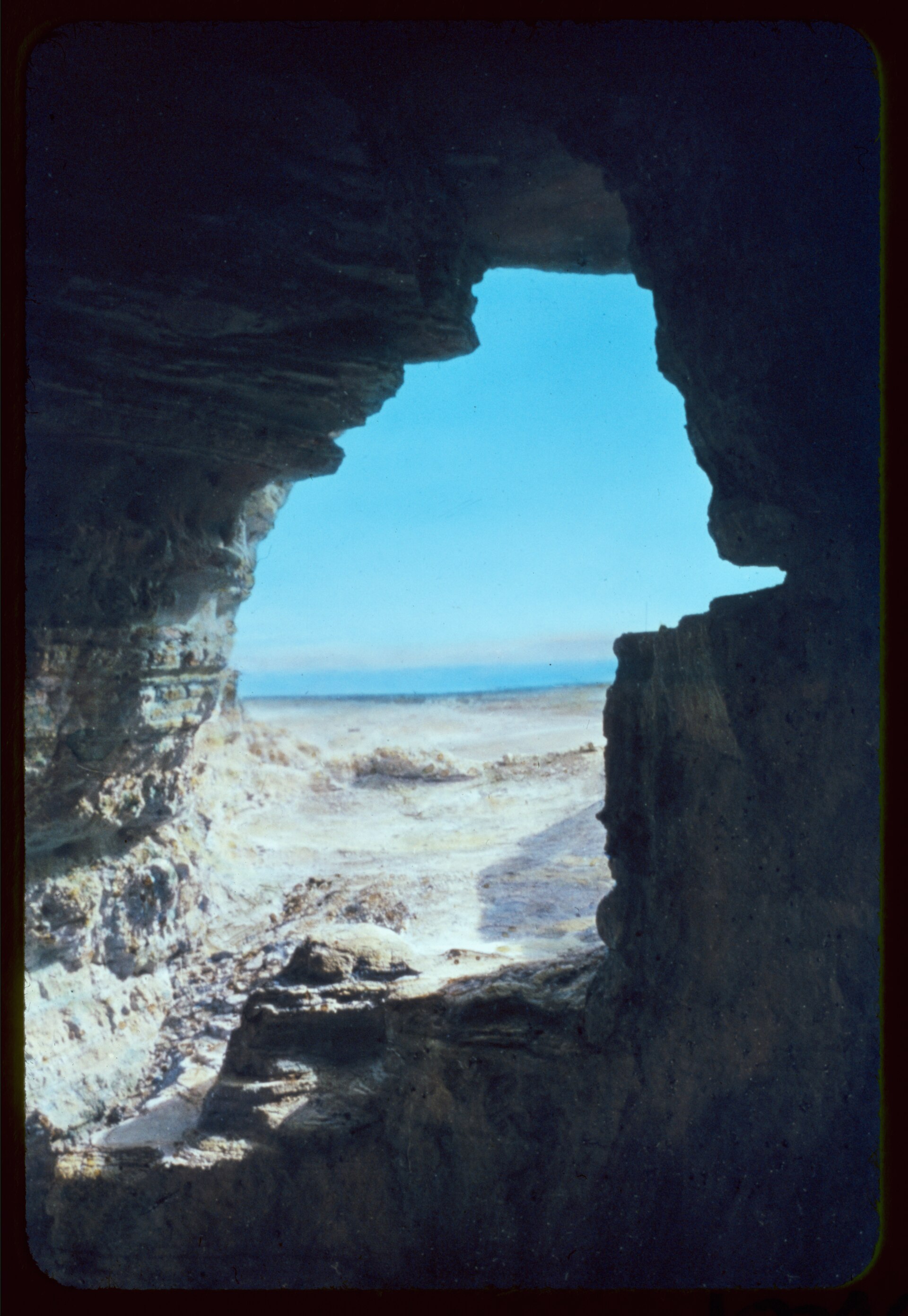 Black and white photograph of steep eroded cliffs and ridges with dark cave openings, and a flat plain and distant water beyond