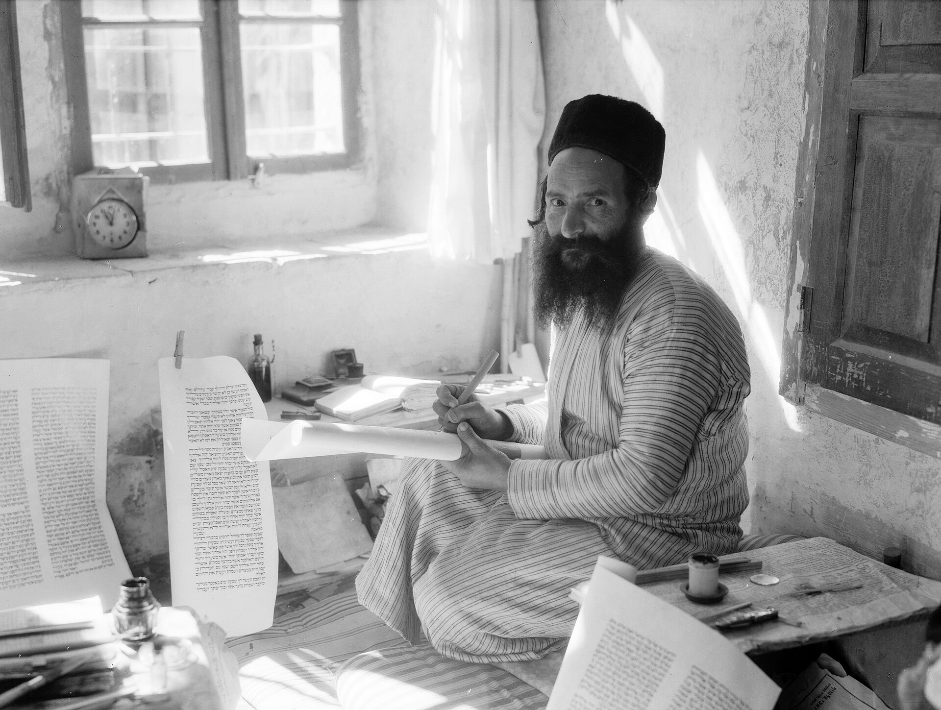 Black and white photograph of a bearded man in a skullcap seated at a table, writing Hebrew letters with a quill on a large sheet of parchment