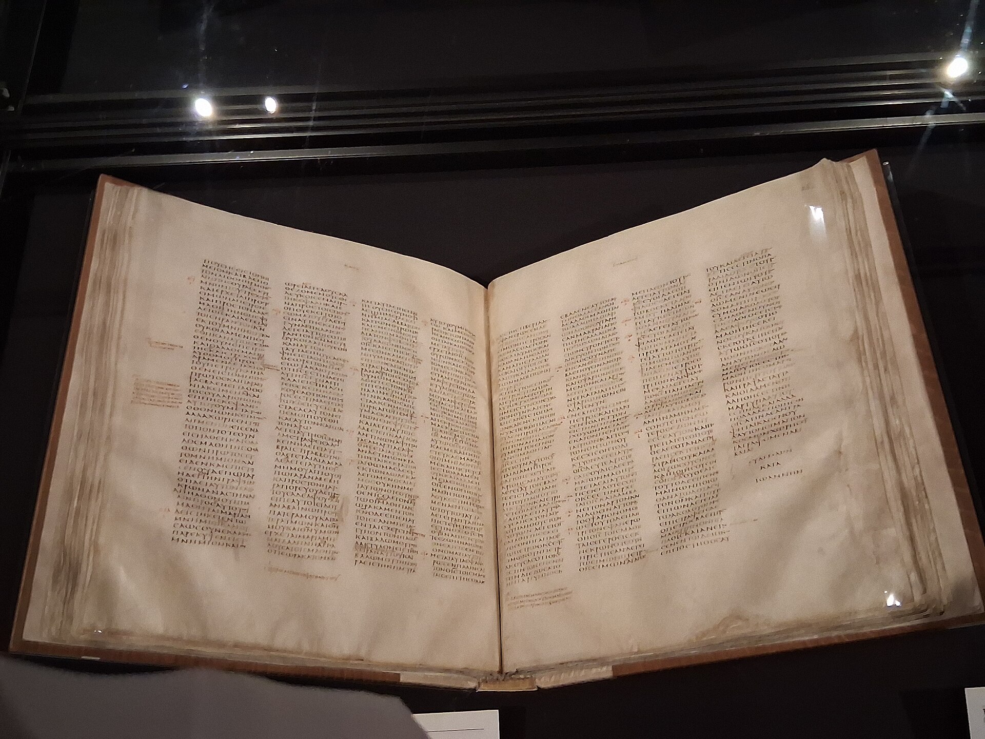An open parchment codex lying in a glass display case, its pages covered in narrow columns of small dark Greek letters