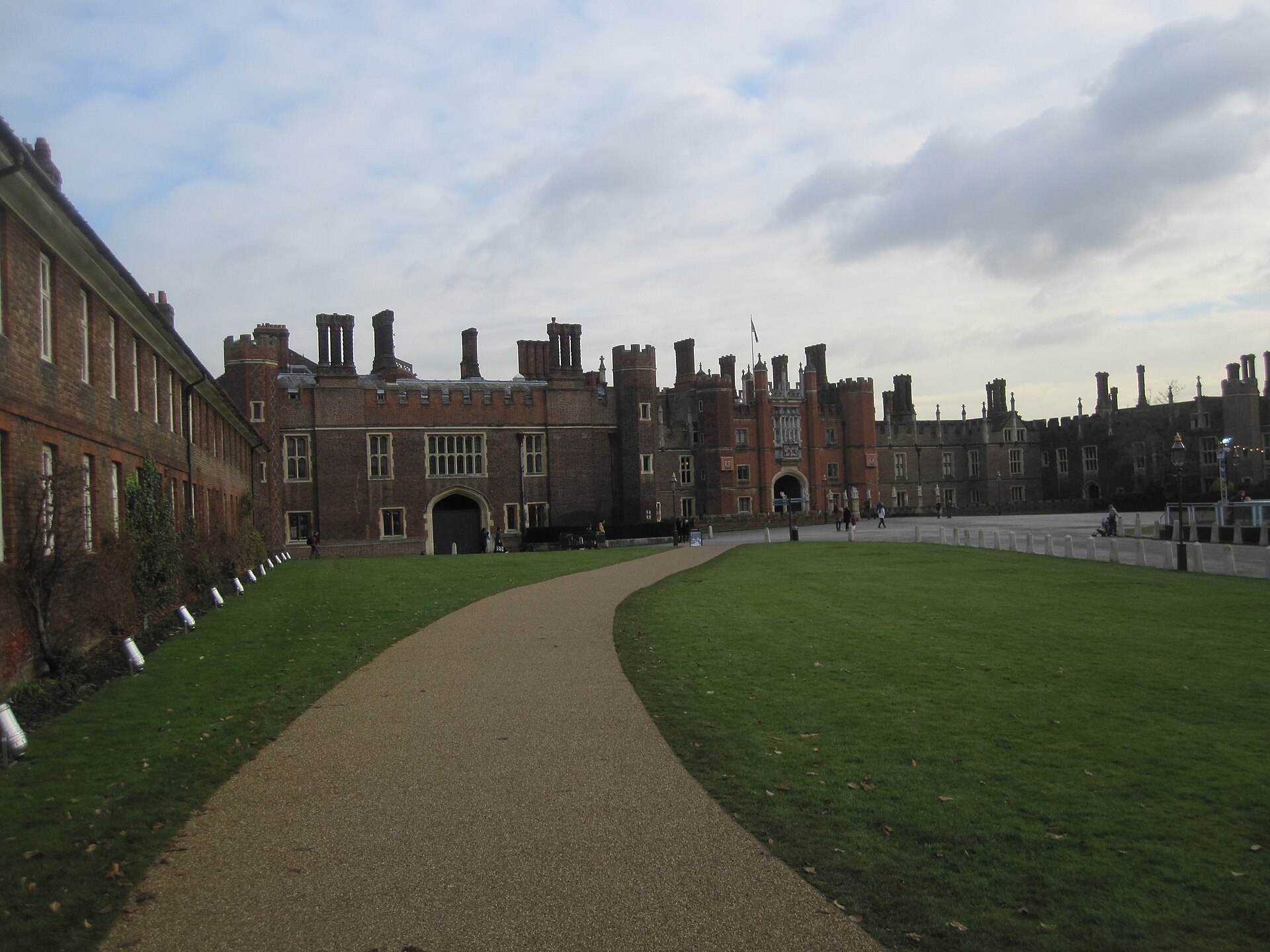 Color photograph of the red brick west front of Hampton Court Palace, with turrets, chimneys and a central gatehouse, seen across a wide forecourt under a grey sky