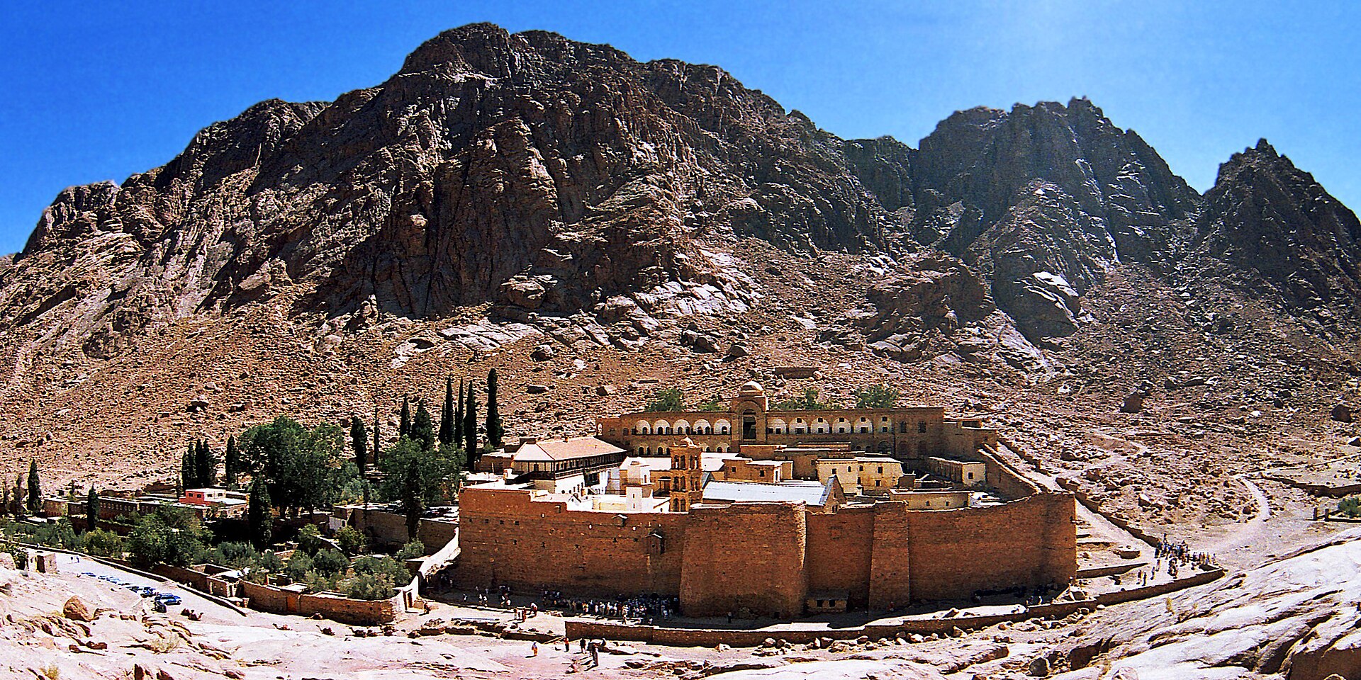 A walled stone monastery with a bell tower and buildings inside the walls, at the foot of steep bare granite mountains