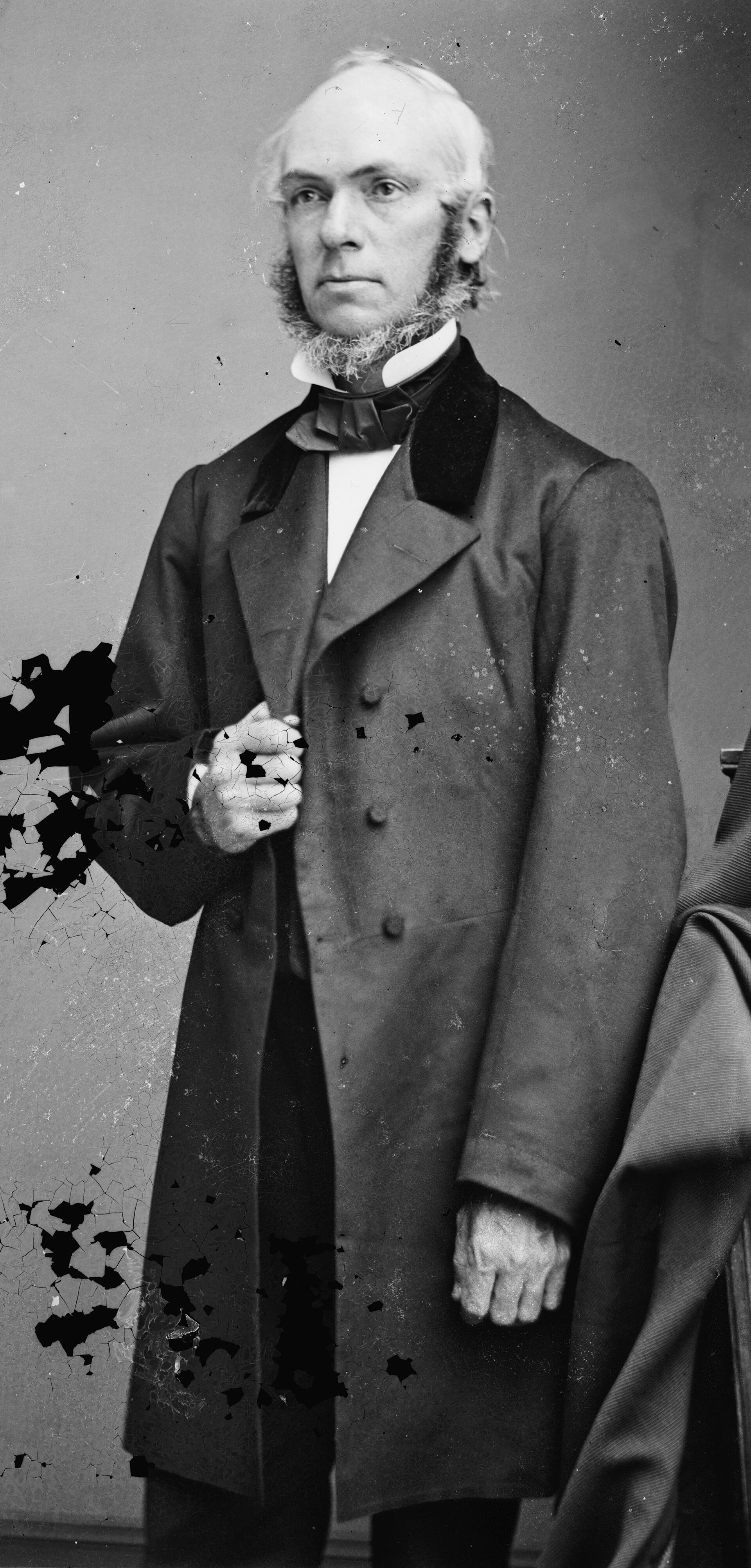 Black and white studio photograph of a bearded man in a dark coat, seated and facing slightly to the side