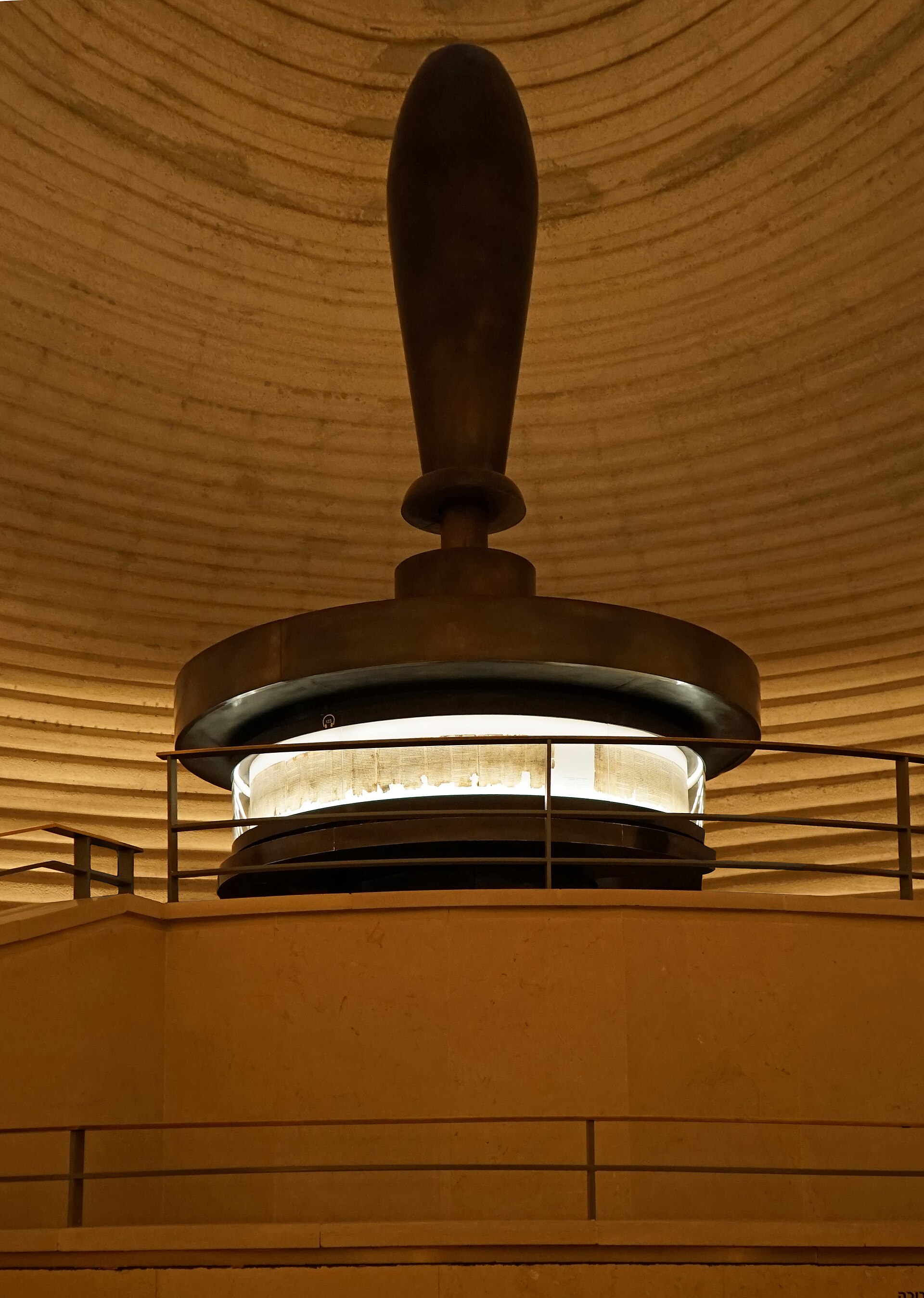 A long Hebrew scroll in columns displayed open in a round glass case under soft light, with visitors' reflections on the glass