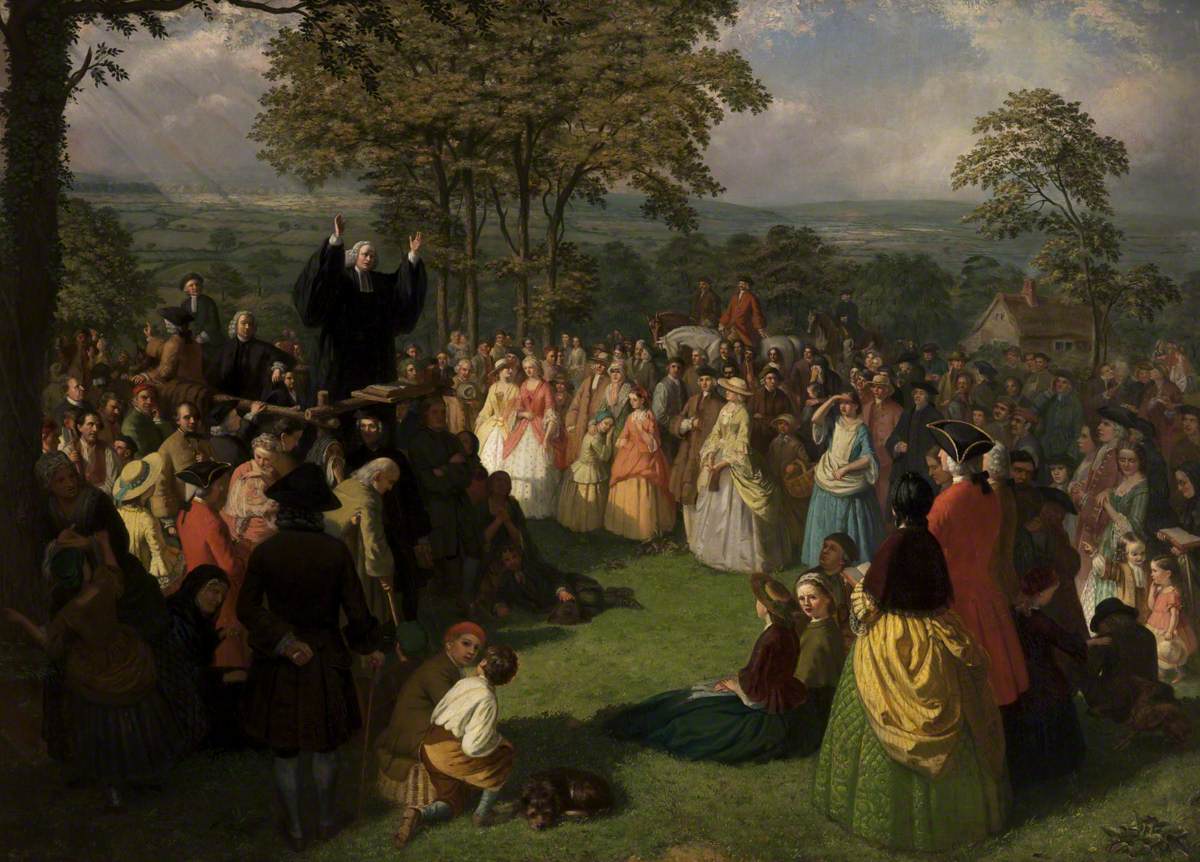 Painting of a crowd of men, women and children gathered in a town street around a preacher in dark clerical dress who stands raised above them with one arm lifted, with buildings and a church tower behind