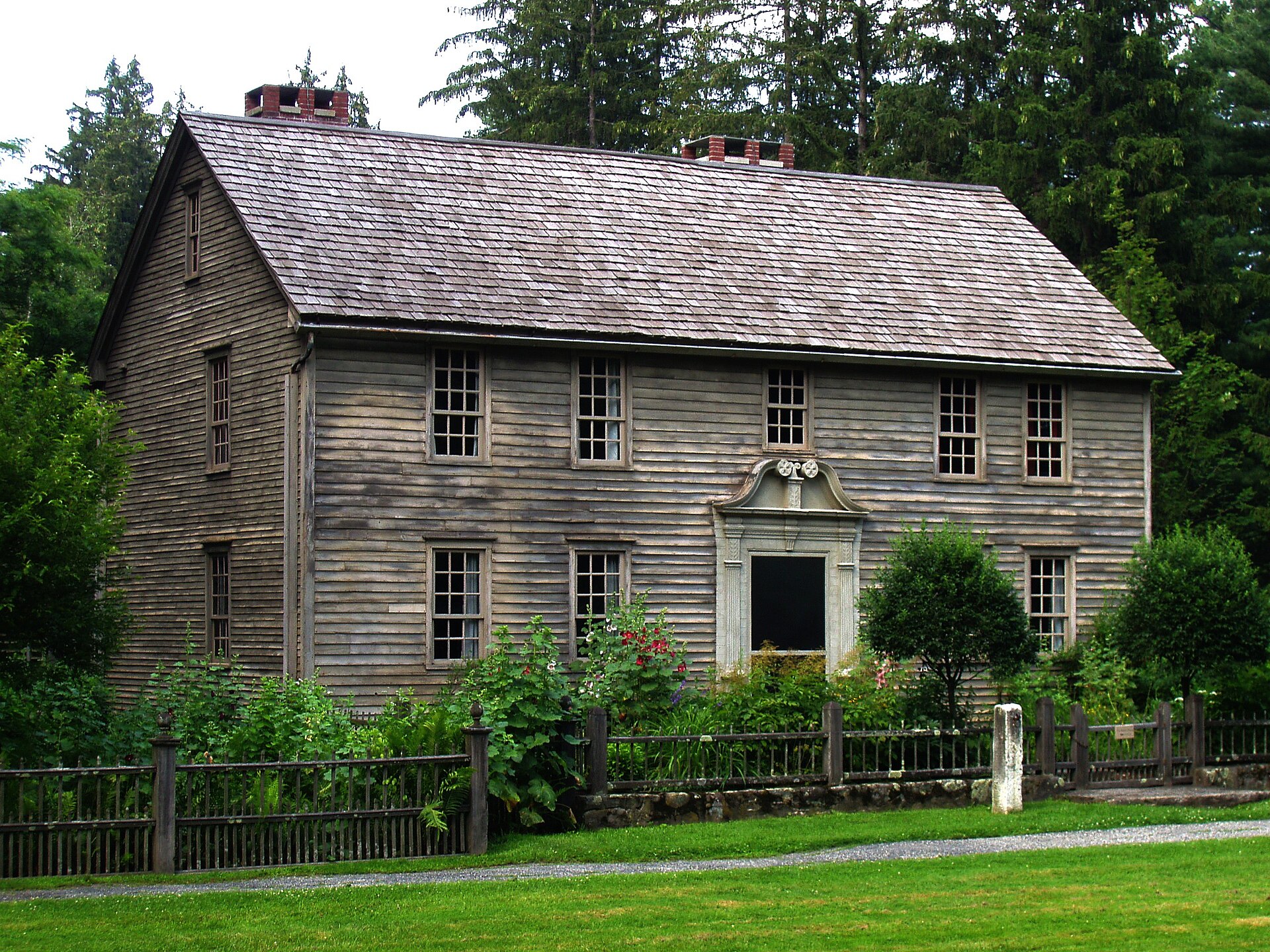 A two-story colonial house painted dark red with a white carved doorway, set behind a lawn and garden on a summer day