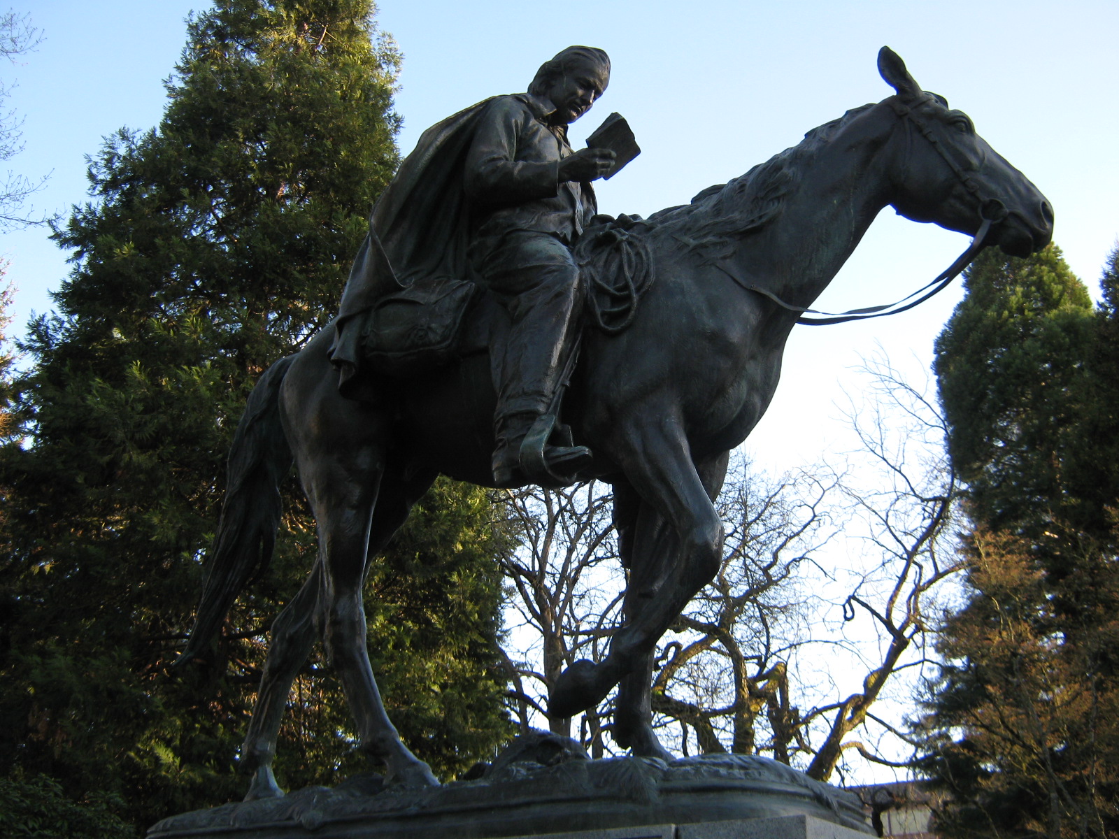 Bronze statue of a man in a broad-brimmed hat and long coat on horseback, reading a book, on a stone pedestal among trees