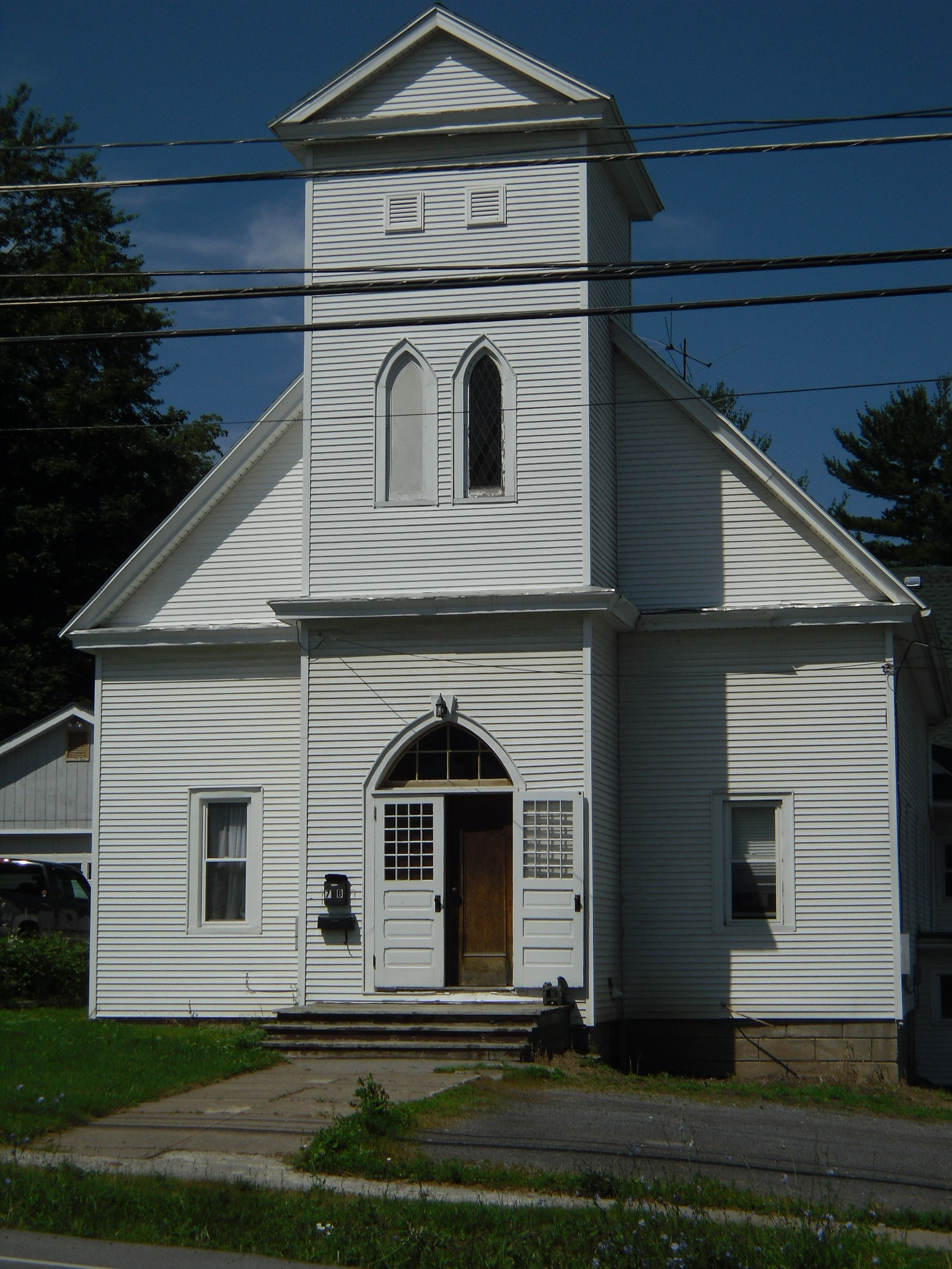 A small-town main street with two- and three-story brick commercial buildings, parked cars and a church steeple in the distance under a blue sky