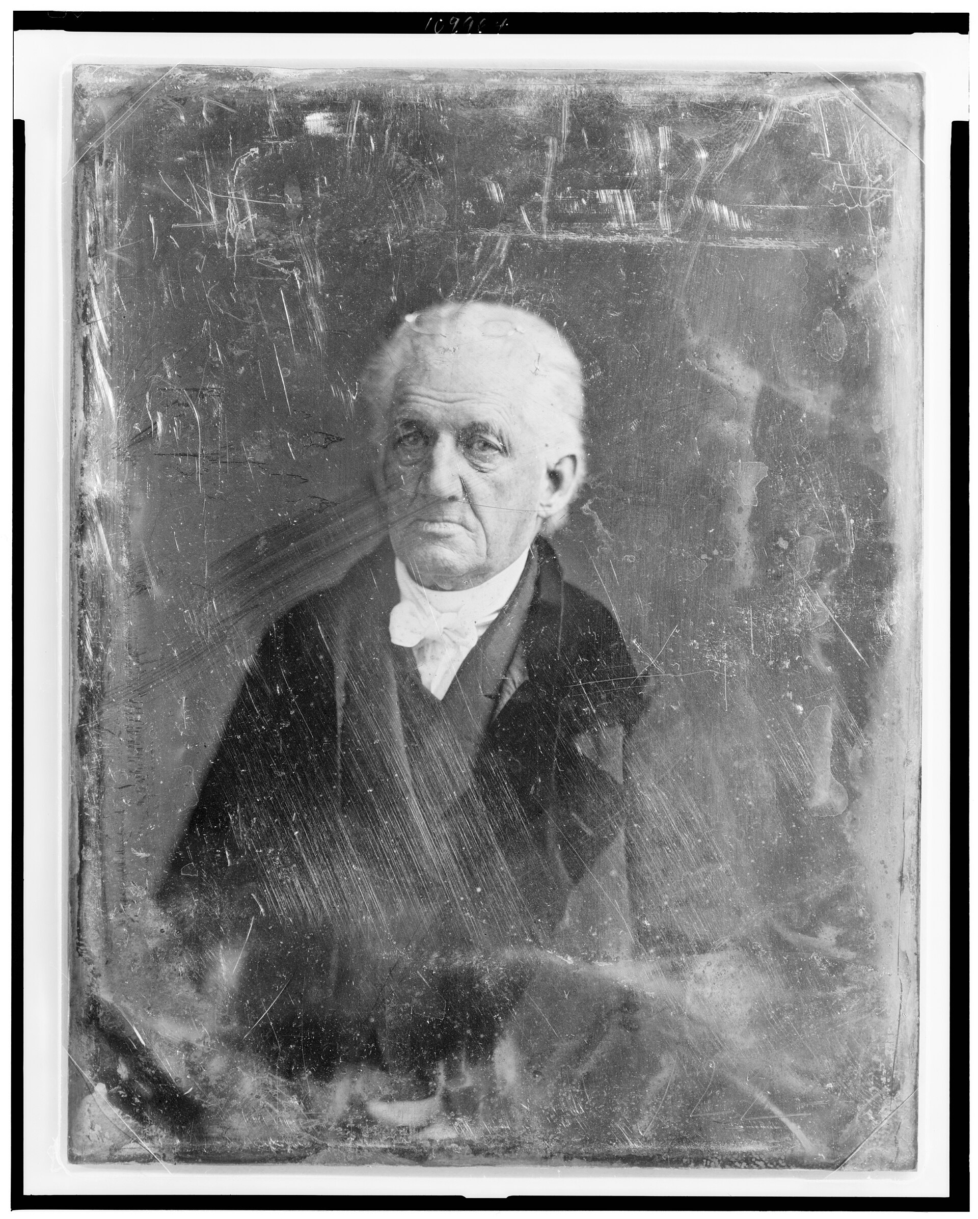 Photograph of an elderly man with white hair brushed back, wearing a dark coat with a high collar, seated and facing the camera