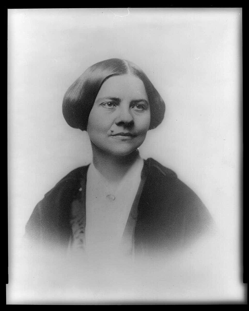 Old photographic portrait of a young woman with dark hair parted in the middle and drawn back, wearing a dark dress with a white collar, facing to the right