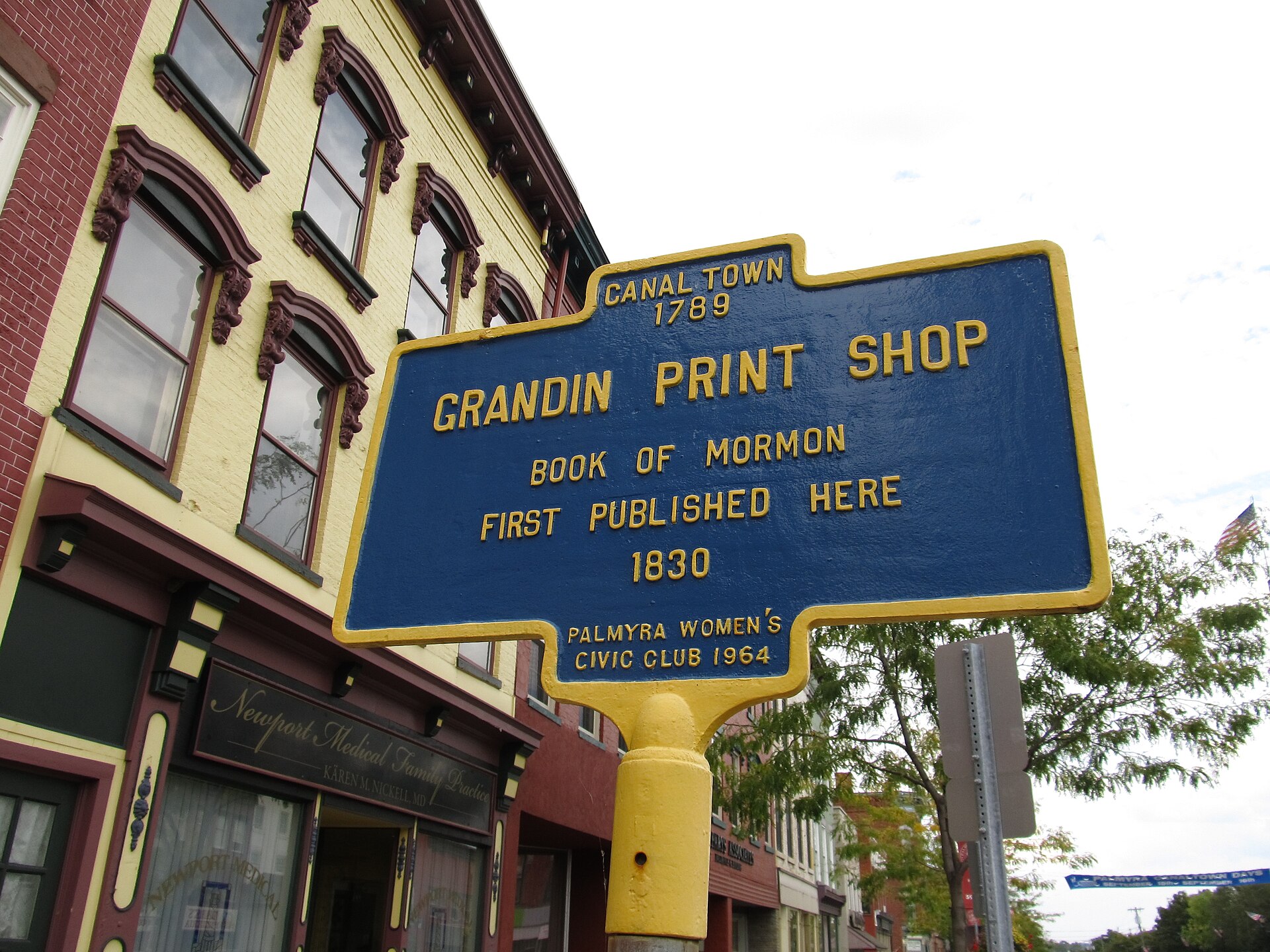 A three-story brick commercial building on a village main street, with tall windows and a sign identifying it as the Book of Mormon publication site