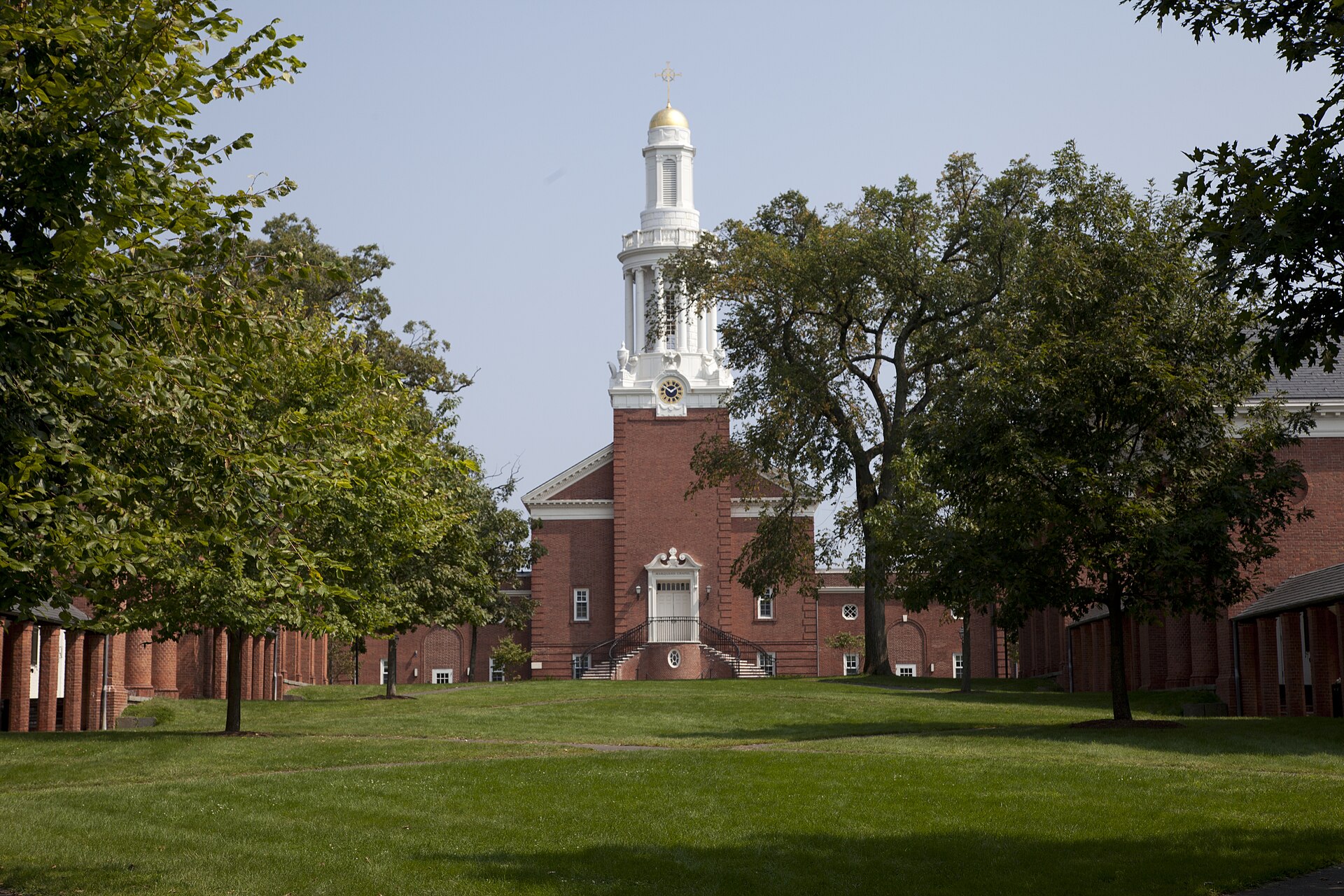 Modern photograph of a quadrangle of red brick buildings with white columns and a white chapel spire, lawn and trees in front, blue sky