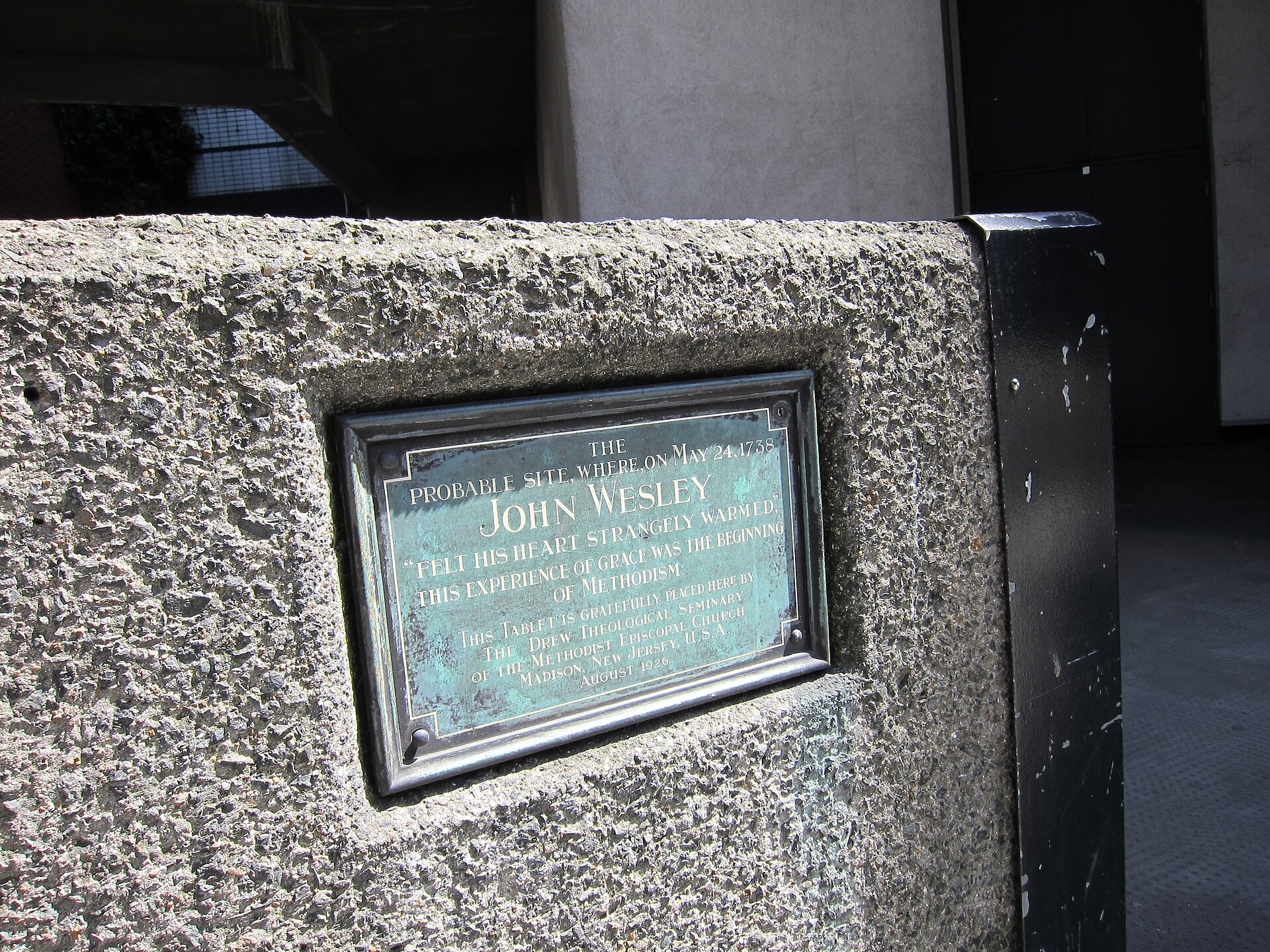 A large bronze plaque mounted on a stone wall in London, shaped like an open book, with raised text quoting John Wesley's journal and an image of flames