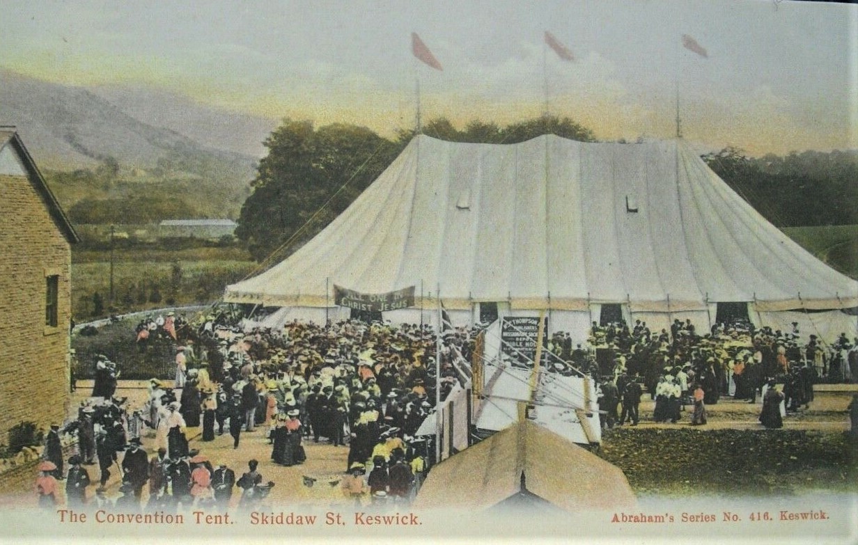 Old black-and-white postcard showing a very large canvas tent with several peaks in a field beside a town street, with trees and hills behind it