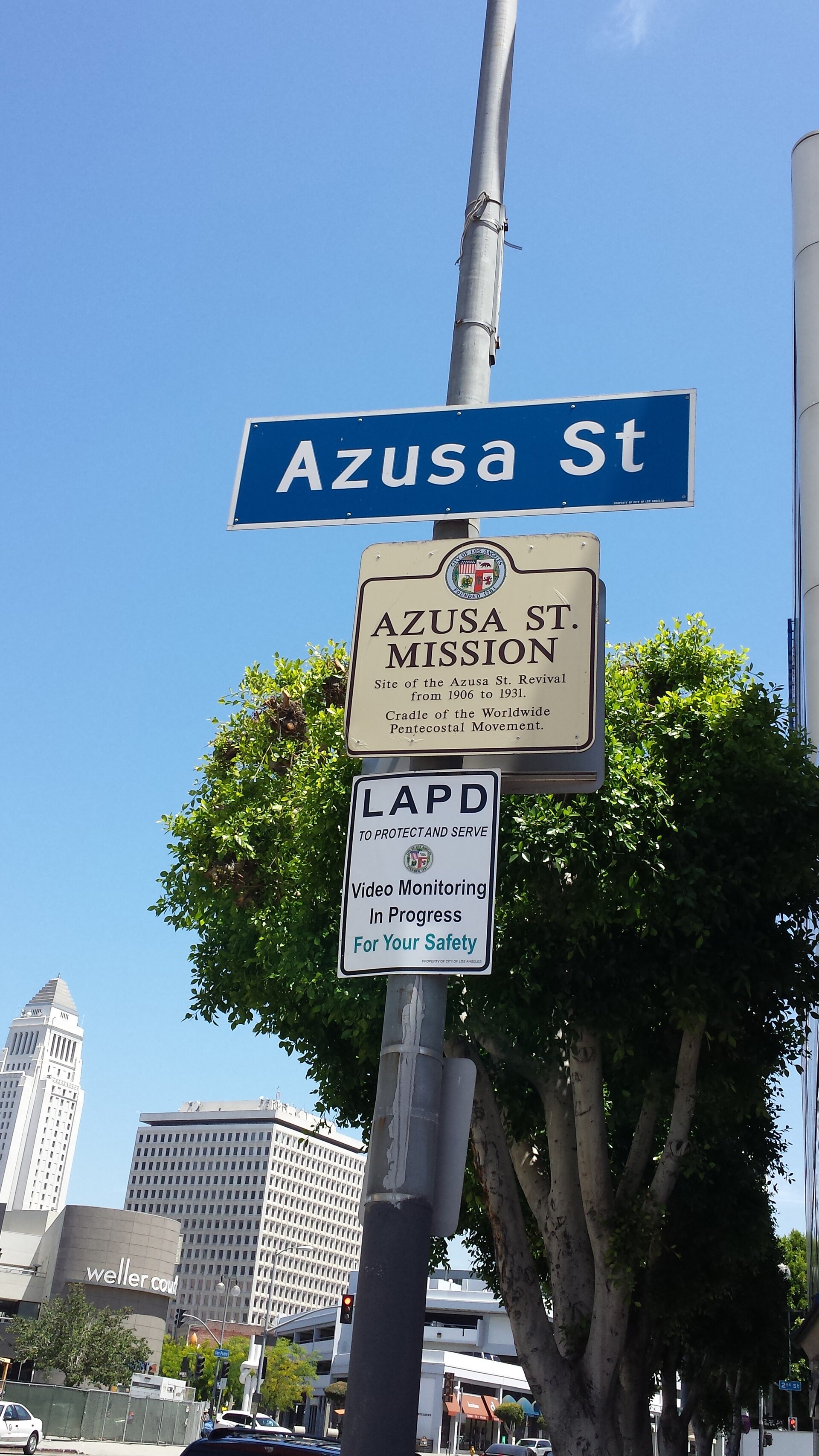 Tall photograph of a historical marker sign mounted on a post in a paved plaza in downtown Los Angeles, with a building behind it