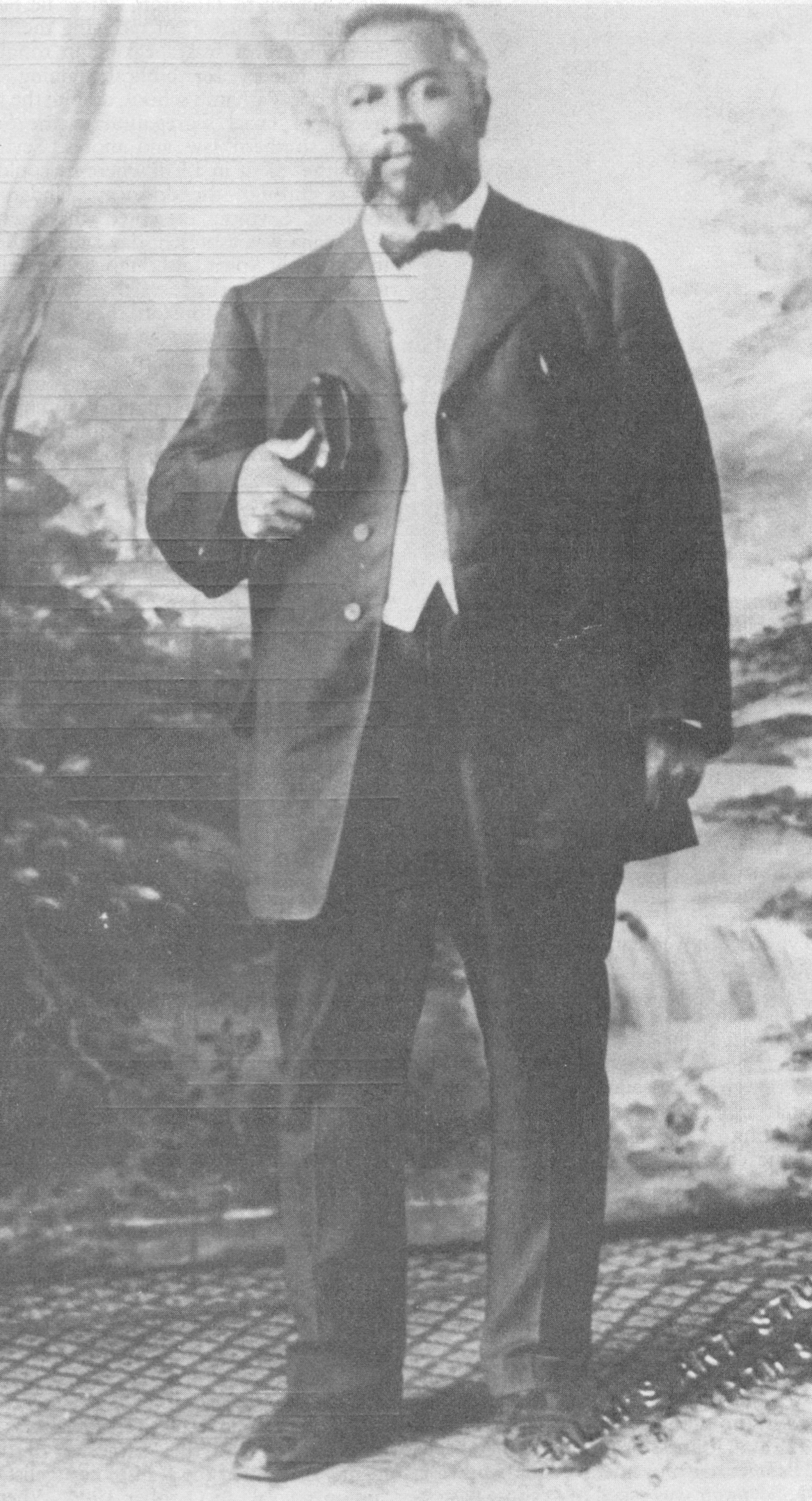 Black and white full length photograph of a Black man with a beard, seated, wearing a dark suit and holding a book on his lap