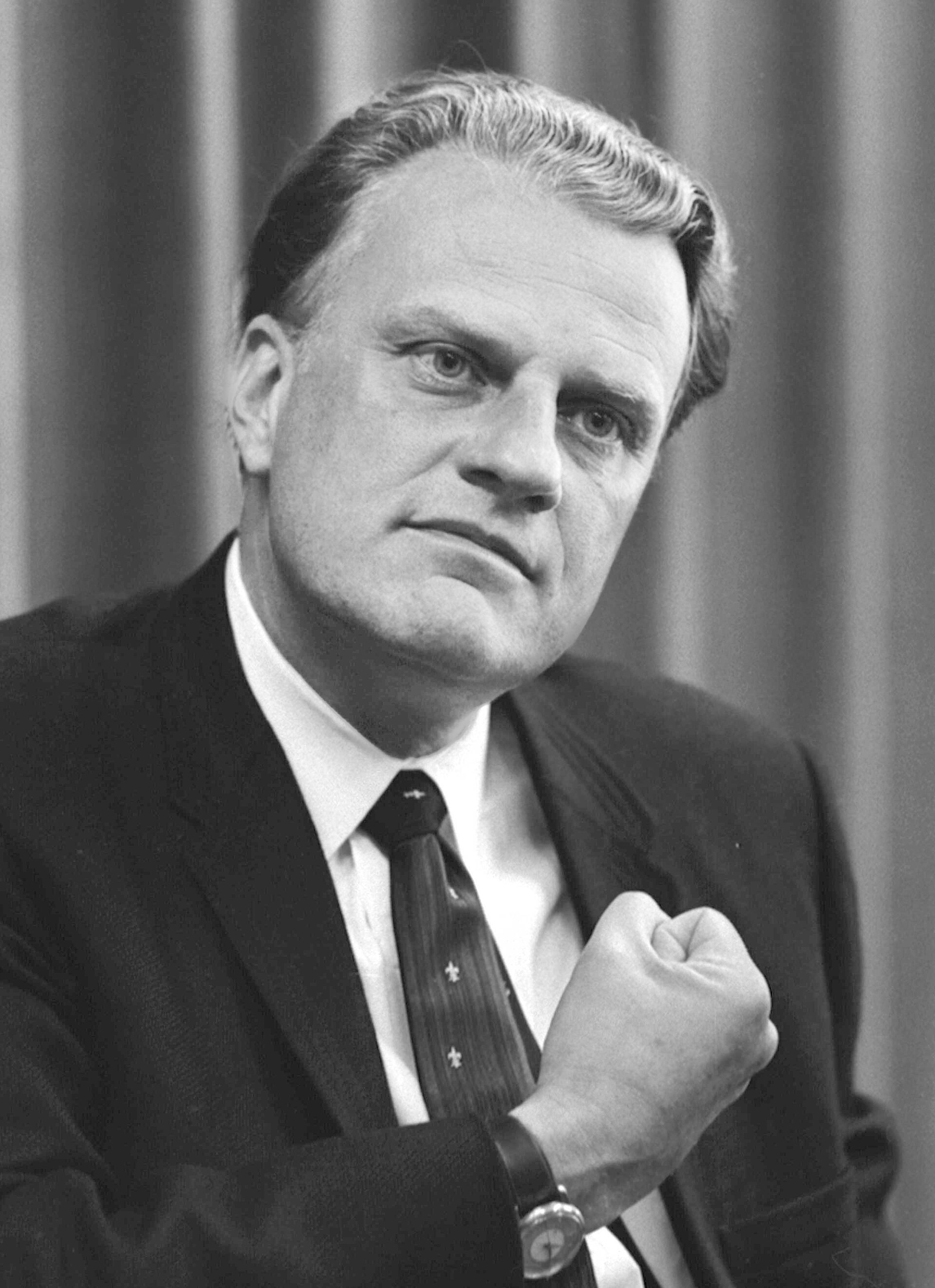 Black and white portrait of Billy Graham in a dark suit and tie, one fist raised to his chest, in front of a curtain