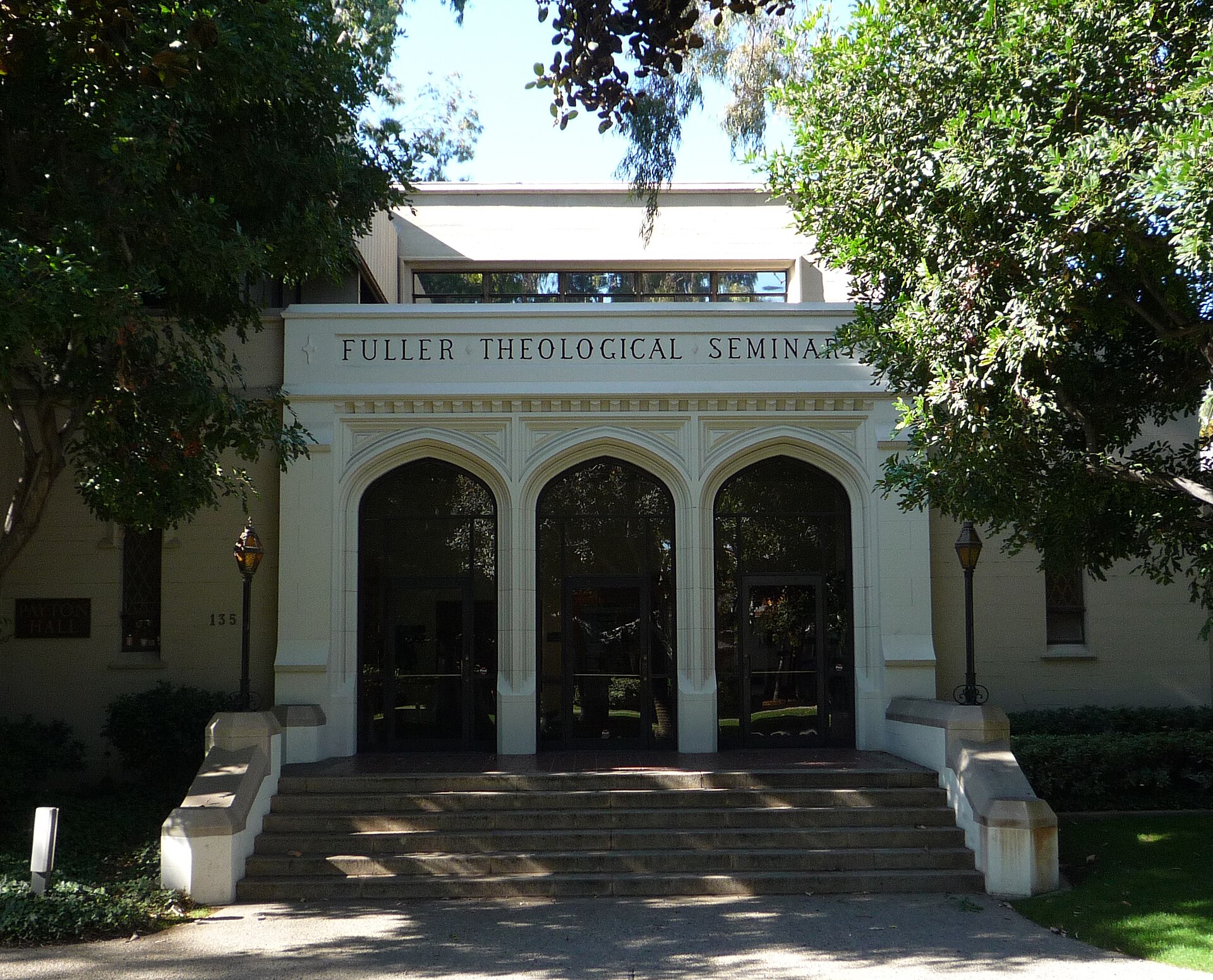 A white building with three arched doorways and the name Fuller Theological Seminary above them, reached by wide steps between trees