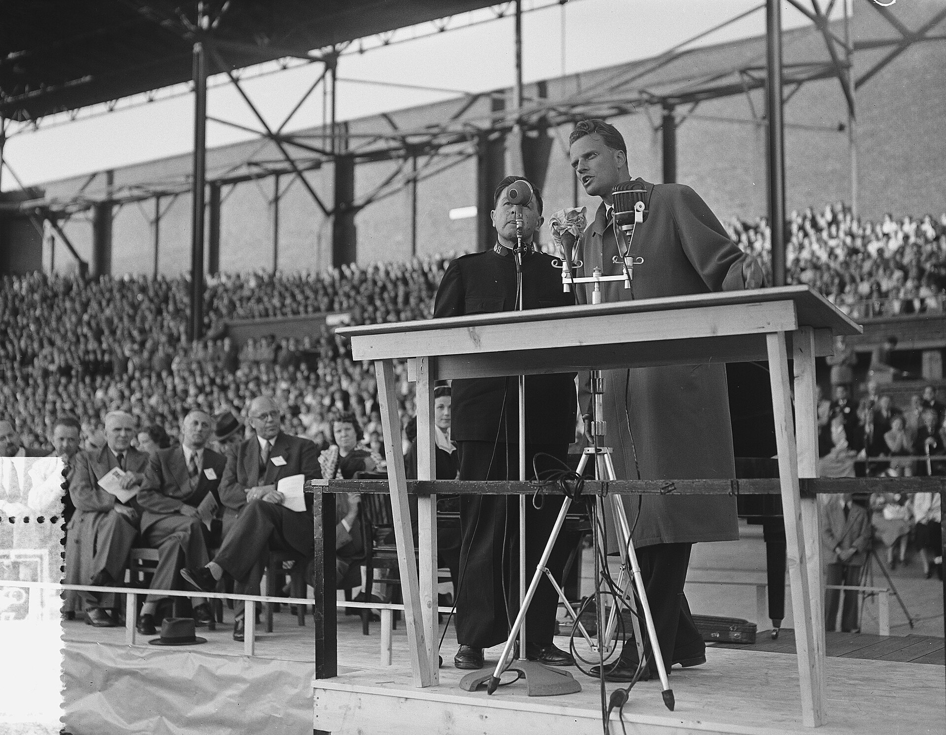 Black and white photograph of Billy Graham speaking at a lectern on a platform with a microphone, a second man standing beside him, and a large crowd seated in covered stadium stands behind them