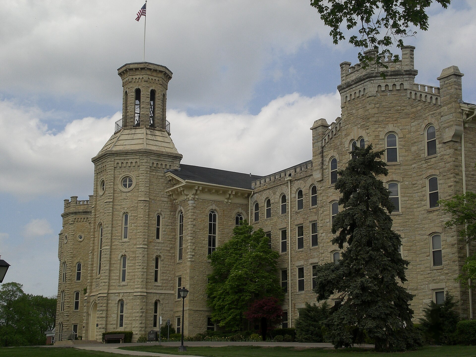 A large limestone building with a tall tower and a smaller turret, with evergreen and leafy trees in front, under a cloudy sky