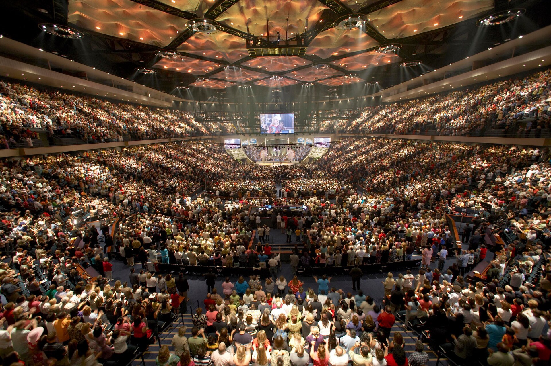 A worship service in a large arena-style sanctuary, with a lit stage under a large screen and thousands of people on the floor and in the stands