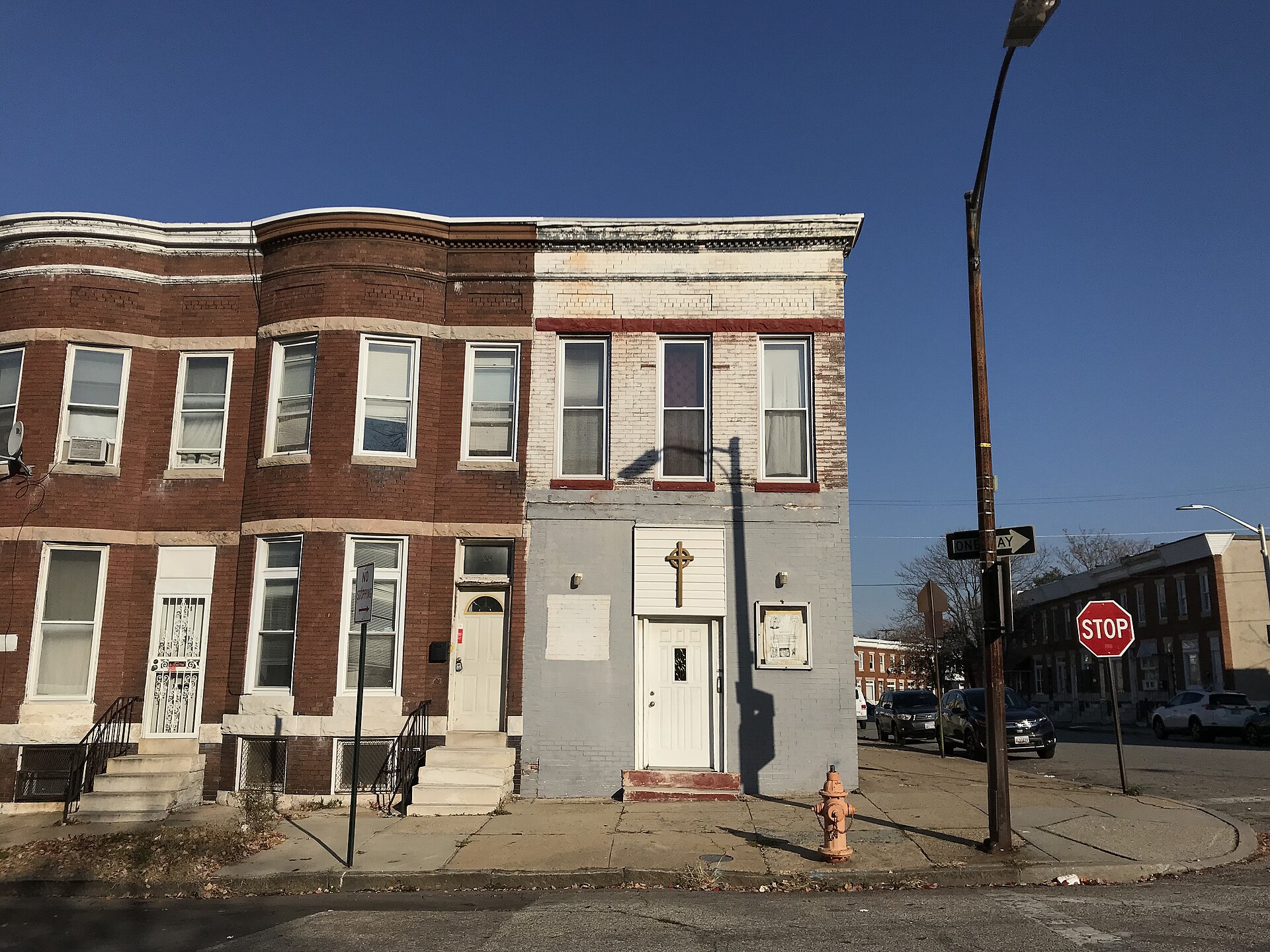 A row of two-story brick buildings on a Baltimore street corner; the gray-painted end building has a cross above its door, with a stop sign and a streetlight at the corner