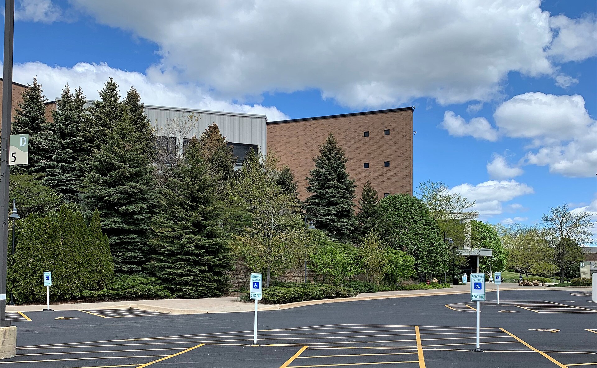 A large parking lot with accessible-parking signs in front of evergreen and leafy trees, with brick and gray church buildings behind them under a blue sky