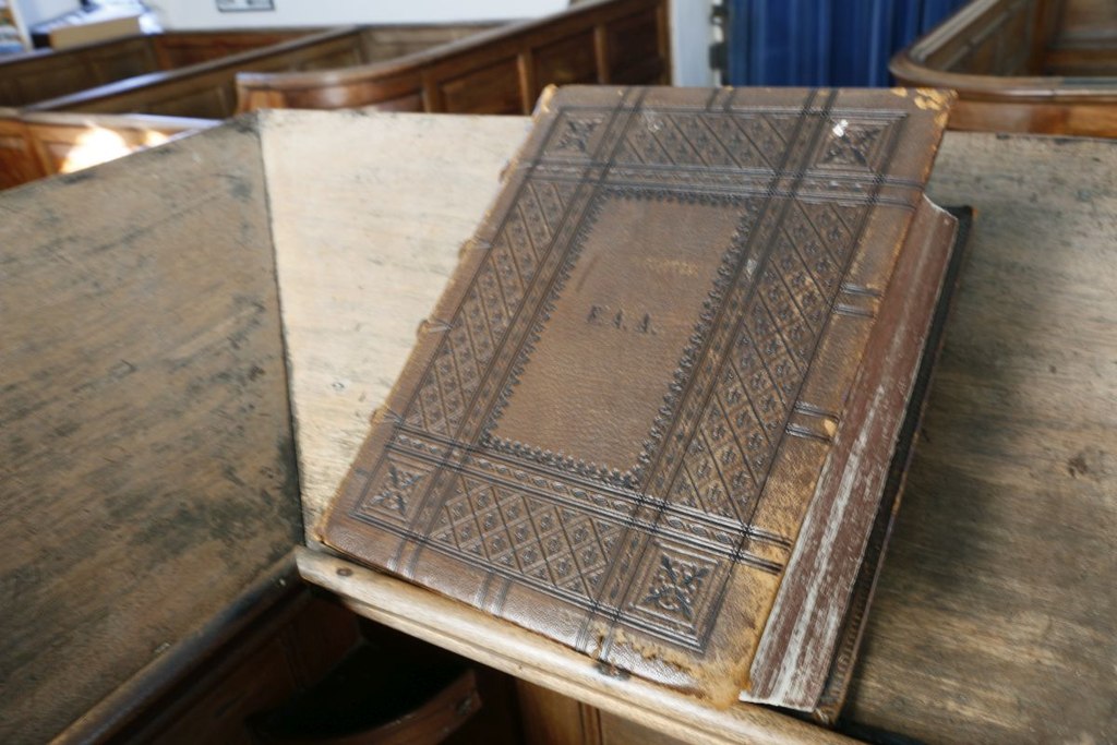A large old Bible with a tooled leather cover lying closed on the slanted top of a wooden pulpit, with wooden pews in the background