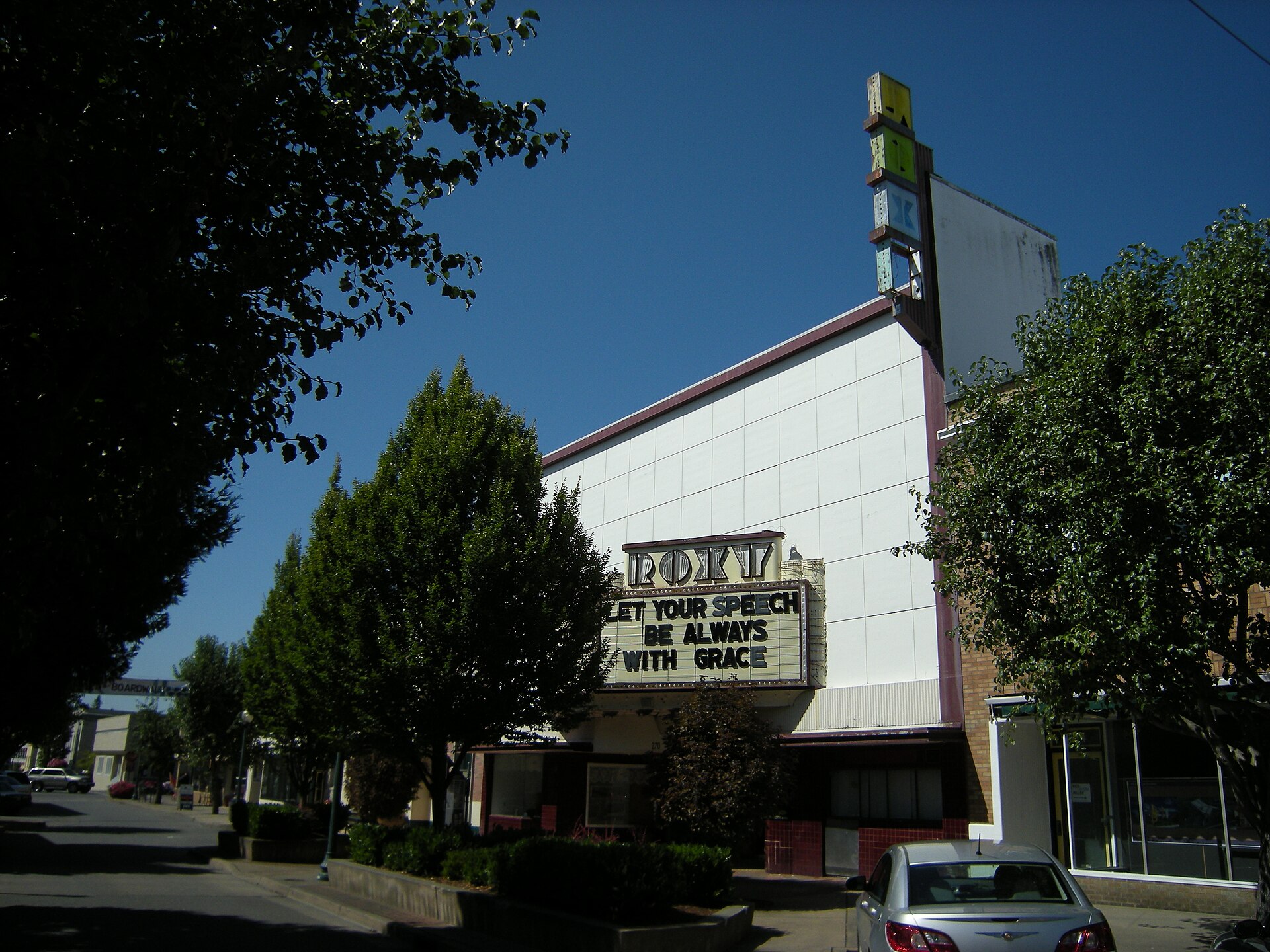 A former movie theater with a tall vertical sign and a marquee reading Let your speech be always with grace, on a tree-lined street under a blue sky