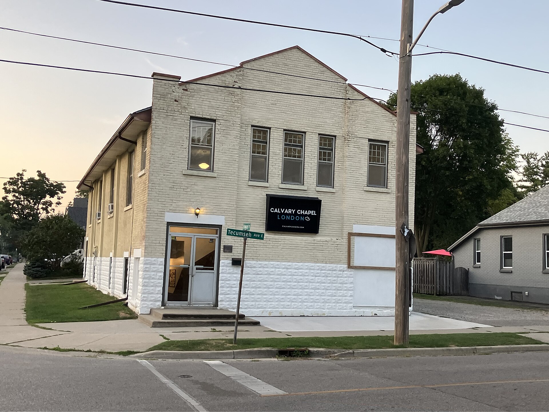 A two-story cream-painted brick building on a street corner, with a black sign reading Calvary Chapel London above the glass front door, a street sign for Tecumseh Ave E, and a utility pole