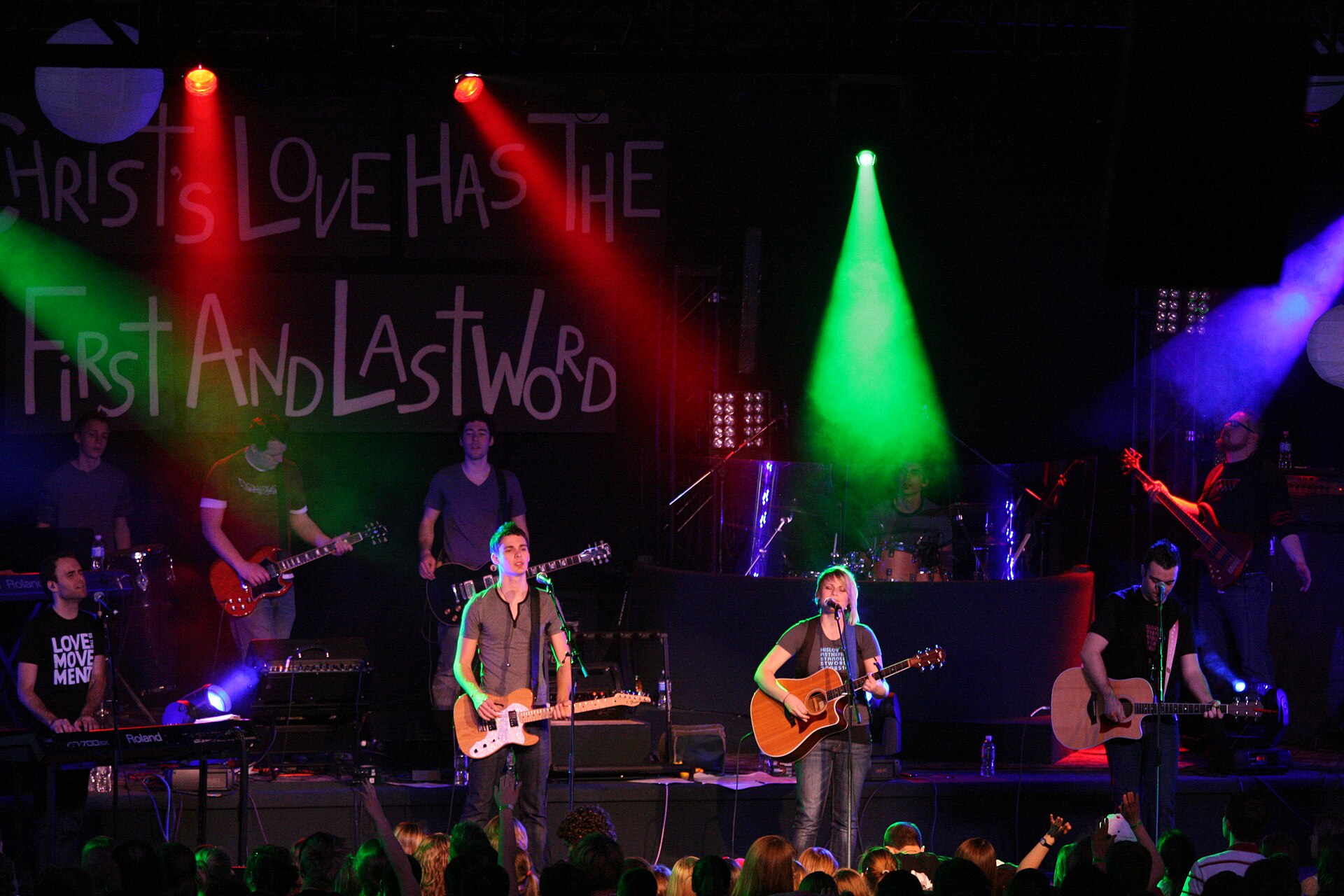 A worship band of guitarists, a keyboard player and a drummer performs on a stage under red, green and blue stage lights; a banner behind them reads Christ's Love Has the First and Last Word, and audience members in the foreground raise their hands.