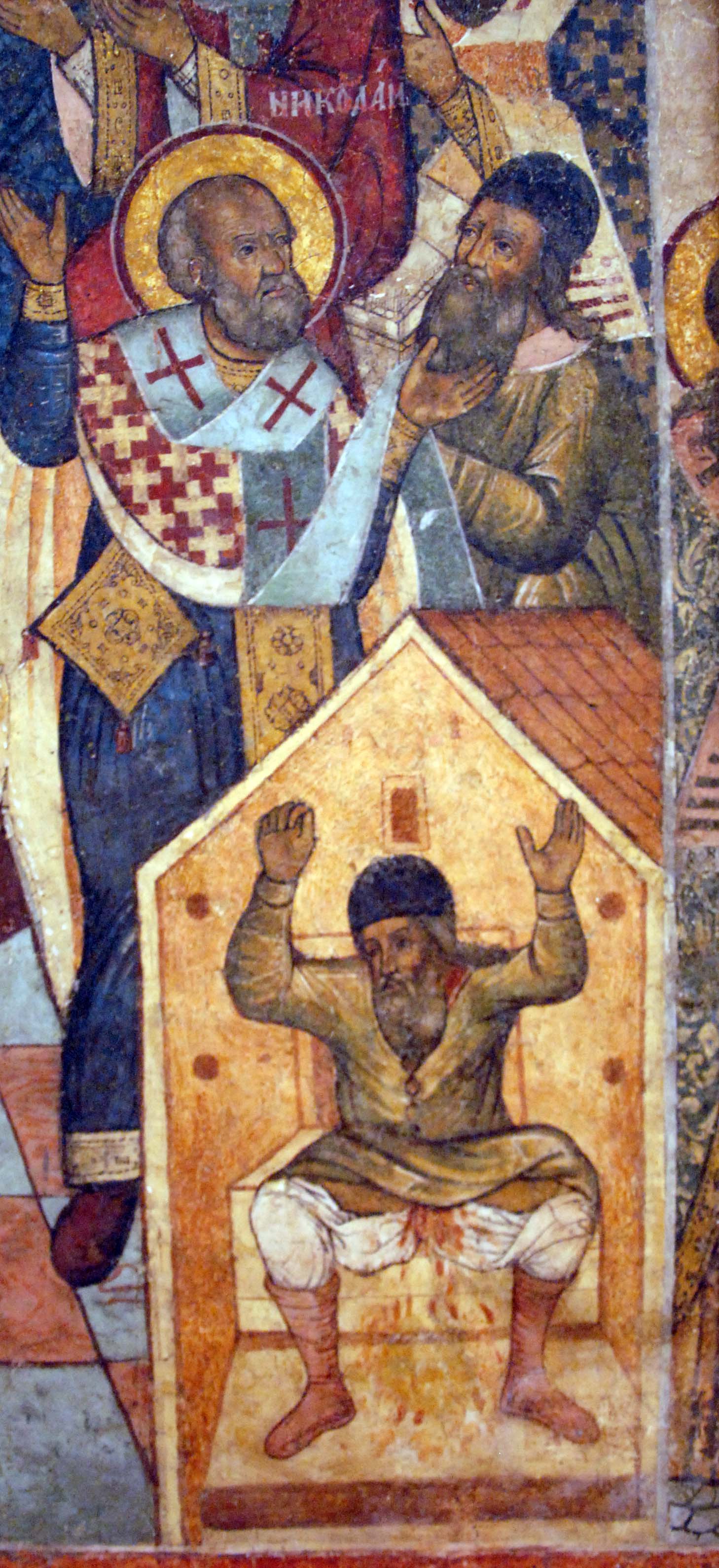 Section of a painted icon: above, a haloed bishop in a cross-patterned vestment raises his hand against the face of a bearded man in a dark cap; below, the same bearded man sits with raised arms inside a small house-shaped structure, with red stains beneath him