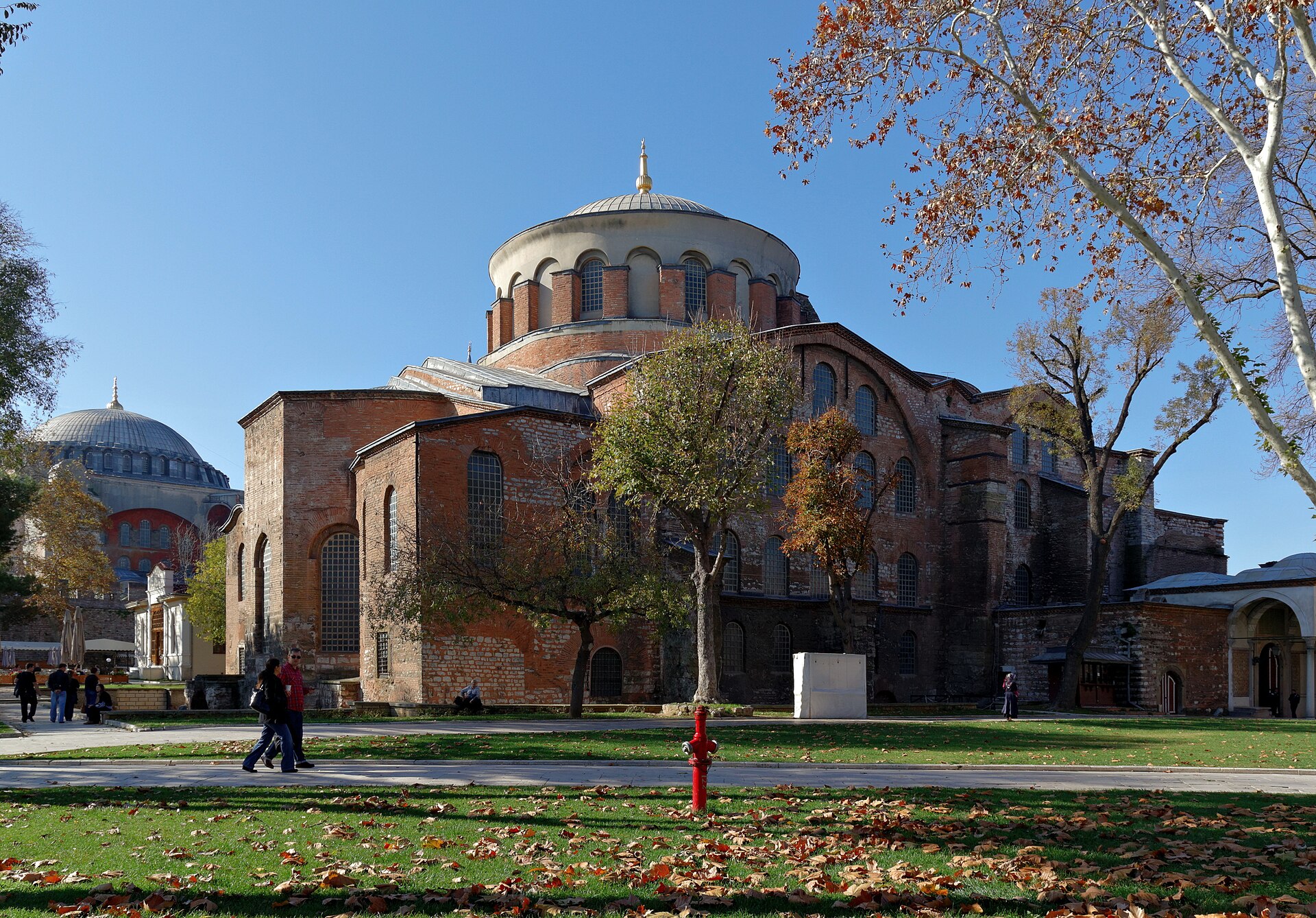 The exterior of a large brick church with a central dome on a drum ringed by windows, smaller half domes and buttresses, and trees in front.