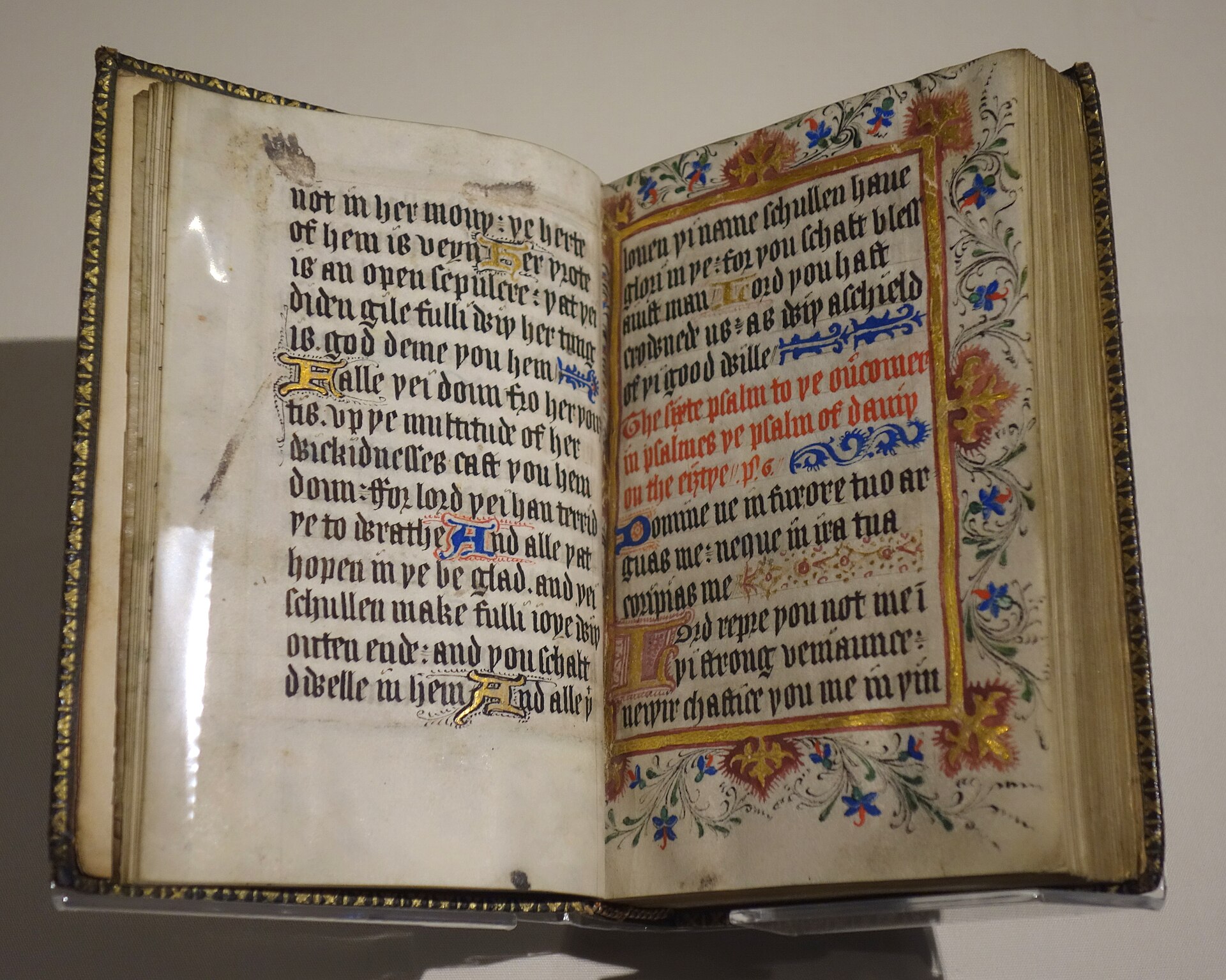 An open handwritten book with two columns of black script, red headings and a decorated initial letter, displayed in a museum case