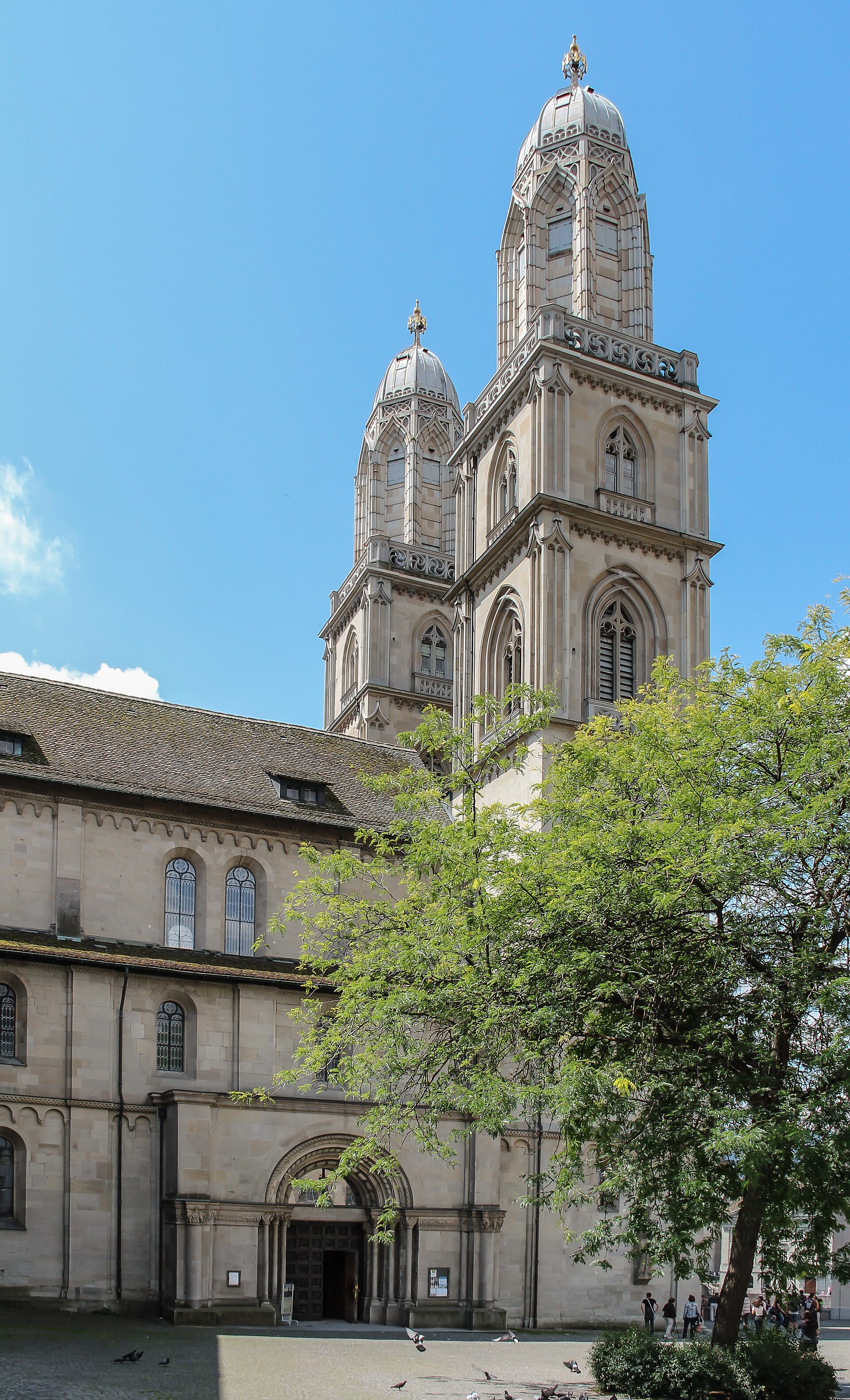 A Romanesque church with two square towers topped by domed caps, seen from below against a blue sky, with houses in front