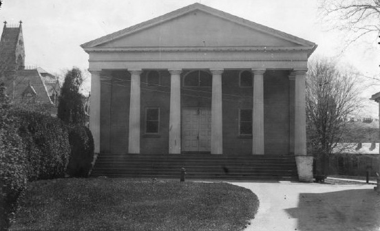 Old photograph of a plain classical chapel building with a columned portico, set among trees beside a larger building