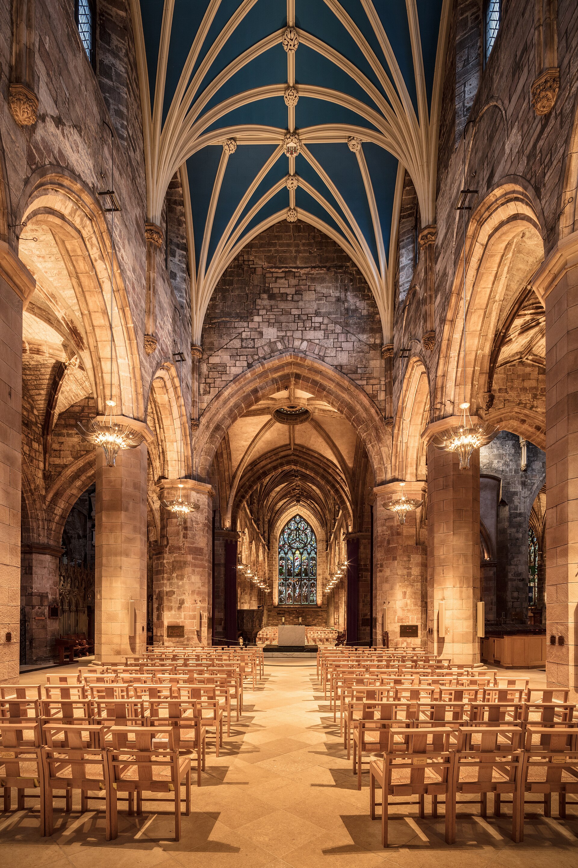 Interior of a stone Gothic church looking down the nave toward a large stained glass window, with clustered pillars, hanging lamps and rows of chairs
