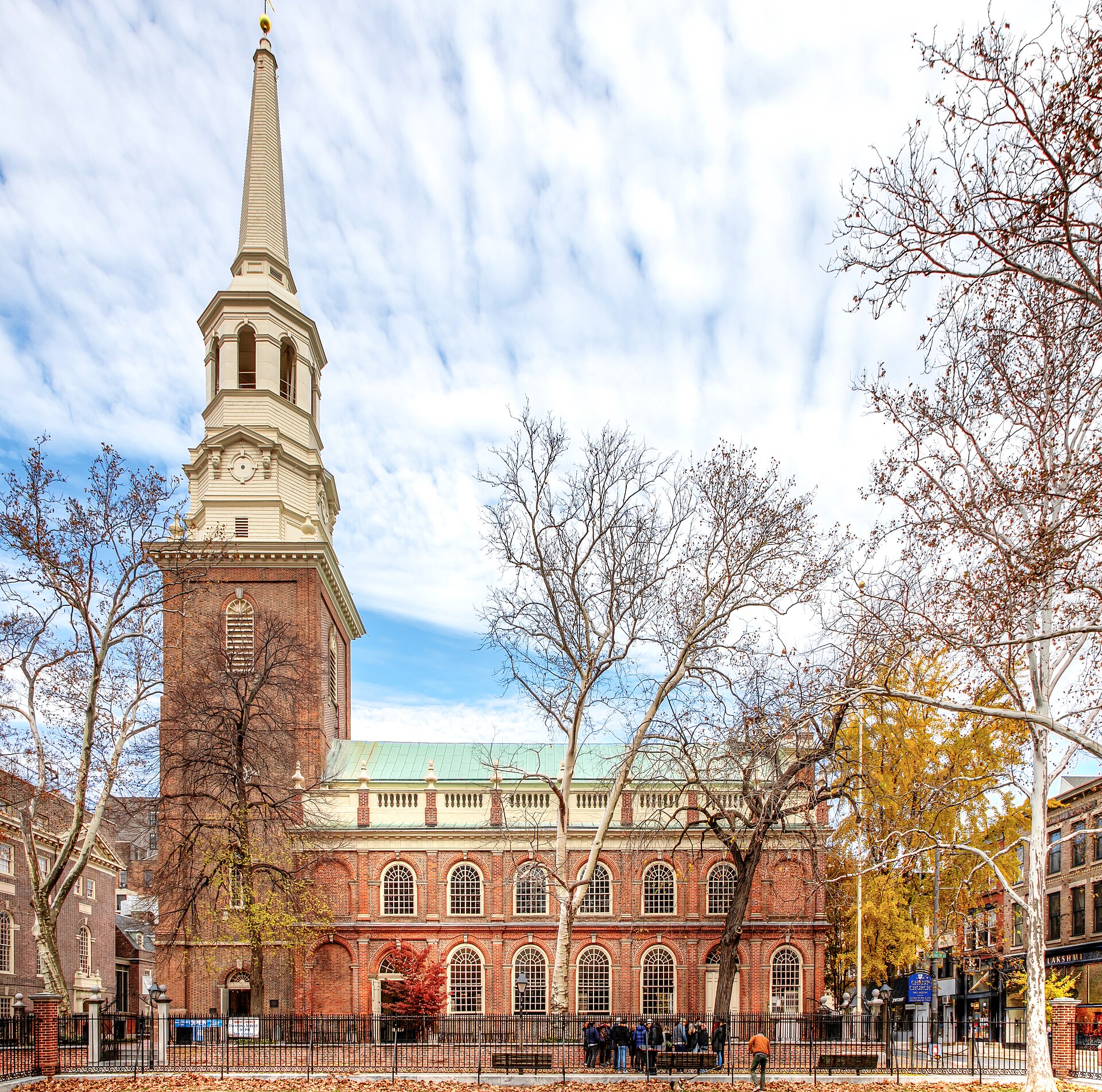 Brick Georgian church with tall arched windows and a white steeple rising in stages to a spire, seen from the street under a clear sky